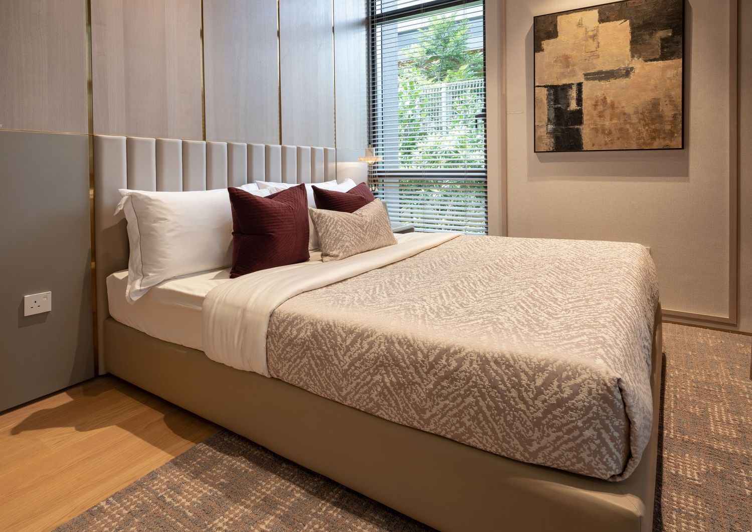 Cozy modern bedroom with beige upholstered bed, burgundy pillows, wood floor, and garden window.