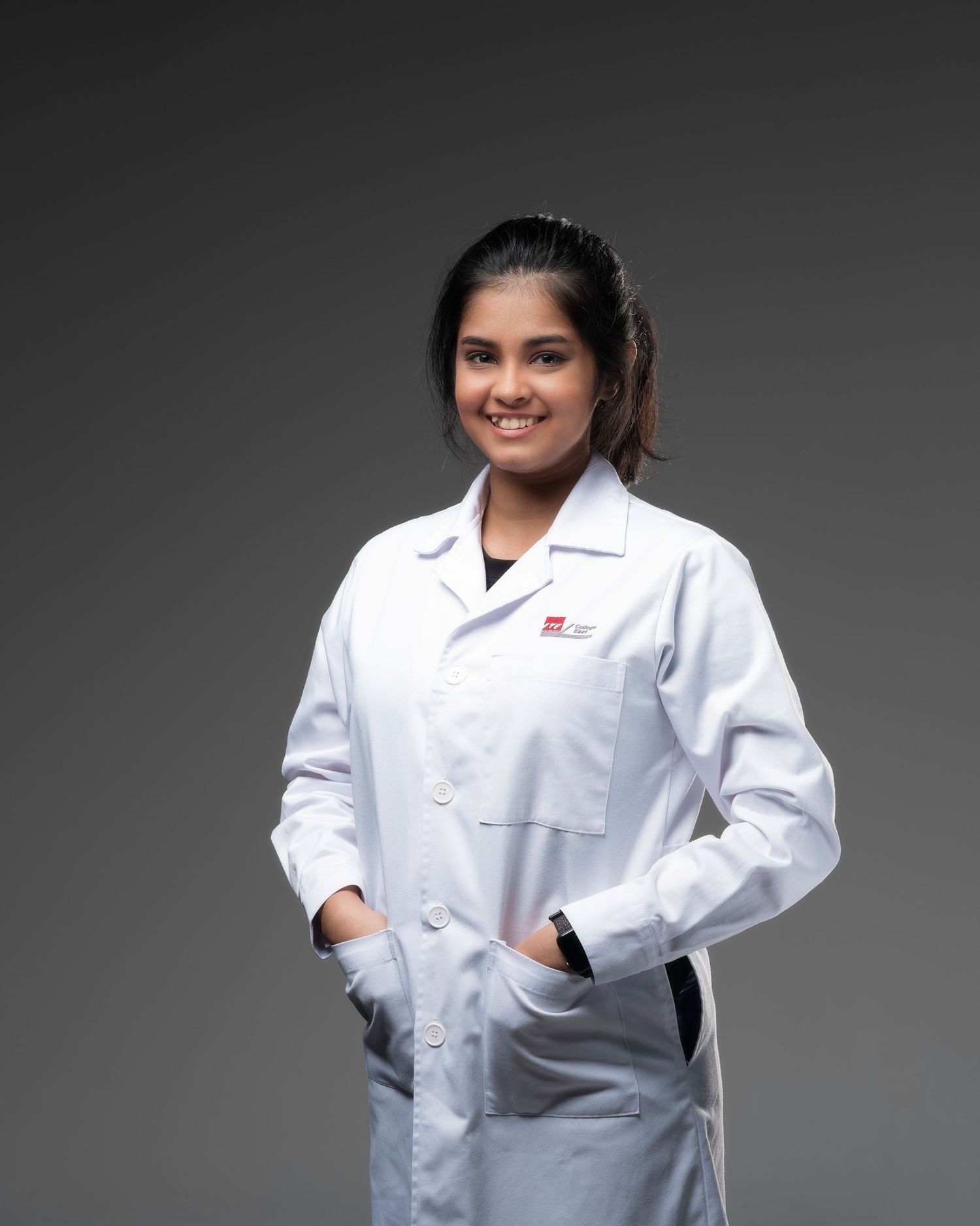 Young woman in white lab coat smiles confidently in a professional portrait against a gray background.