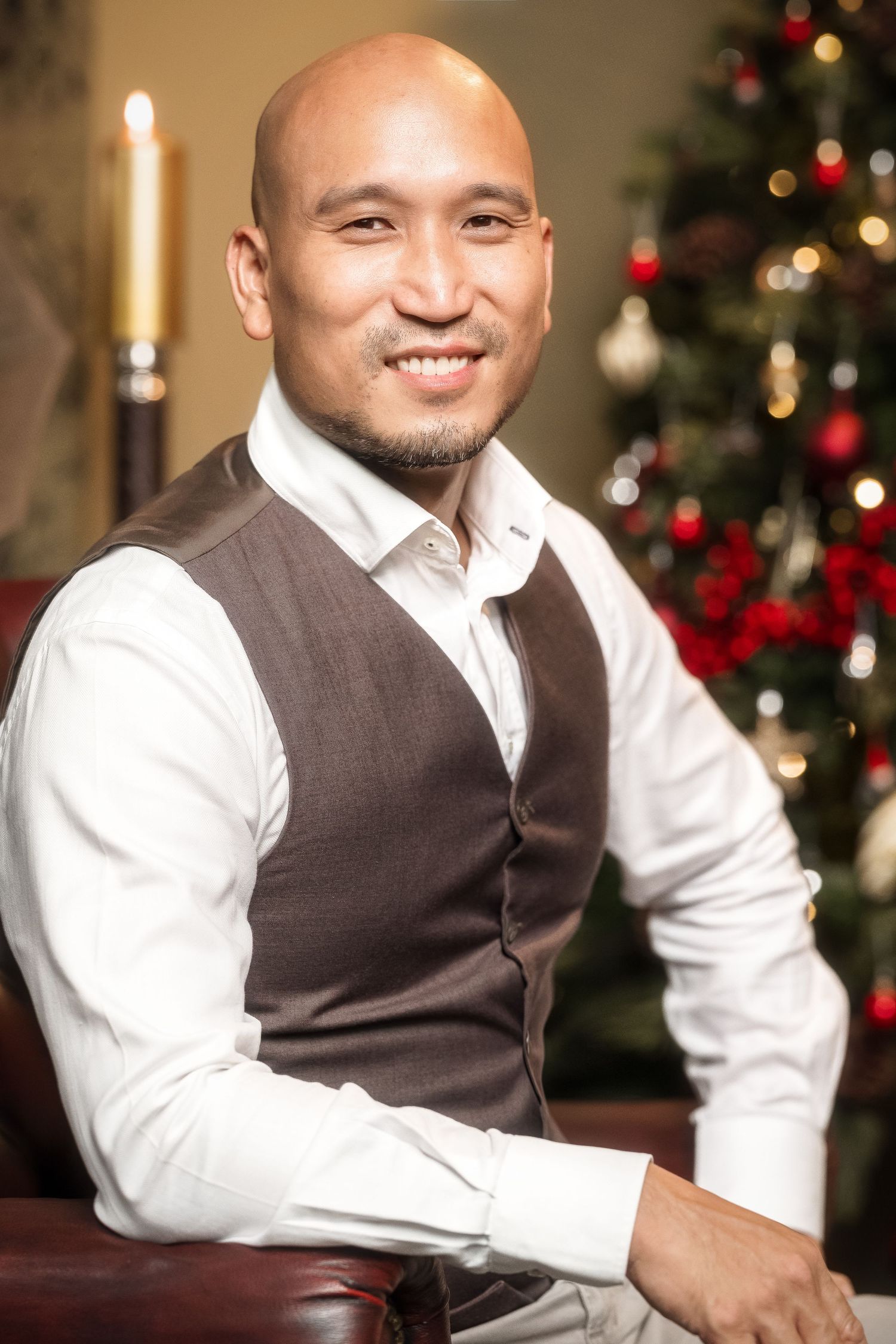 Smiling bald man in vest and white shirt seated near a decorated Christmas tree.