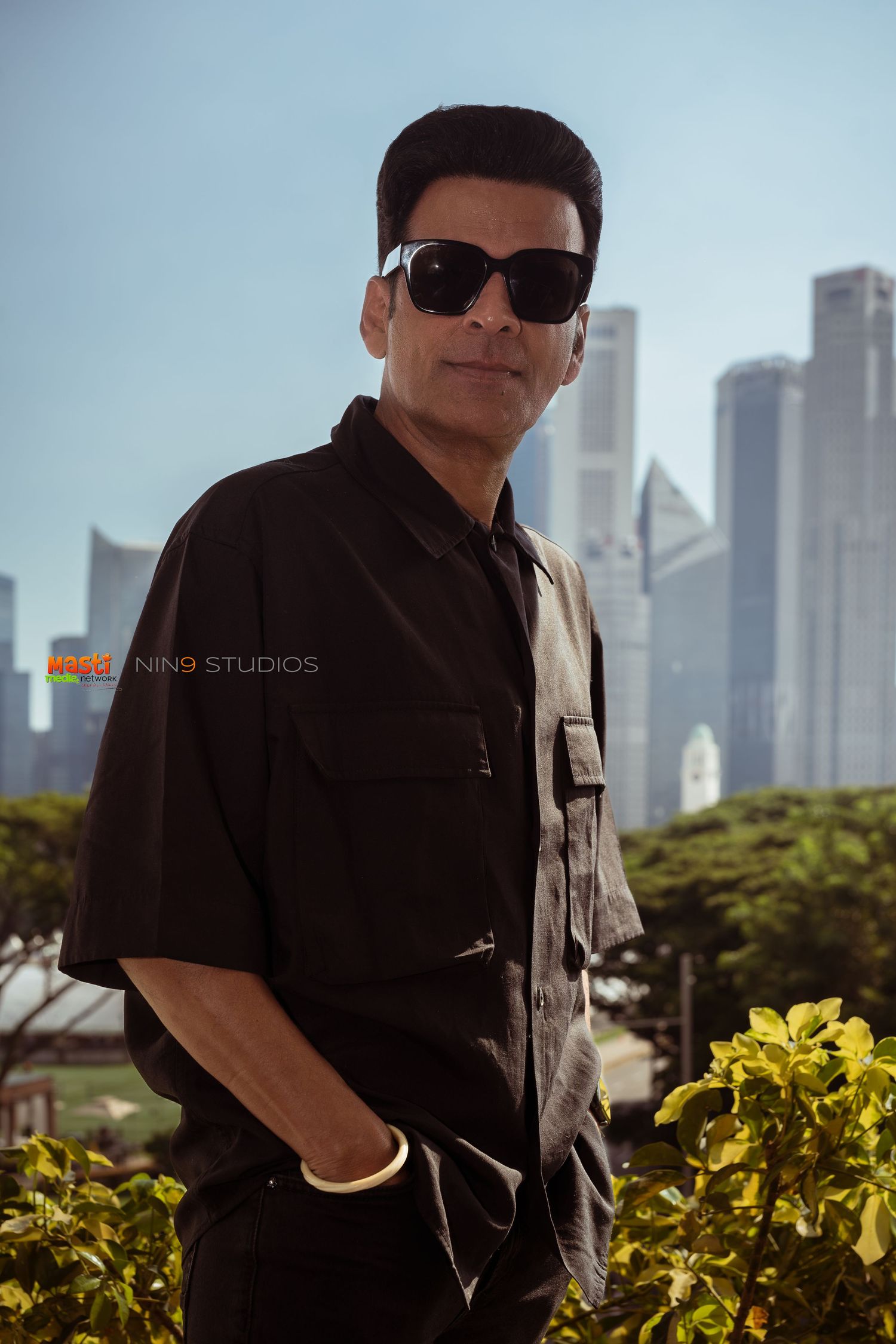 Man in black shirt and sunglasses posing outdoors with city skyline in background on a sunny day.