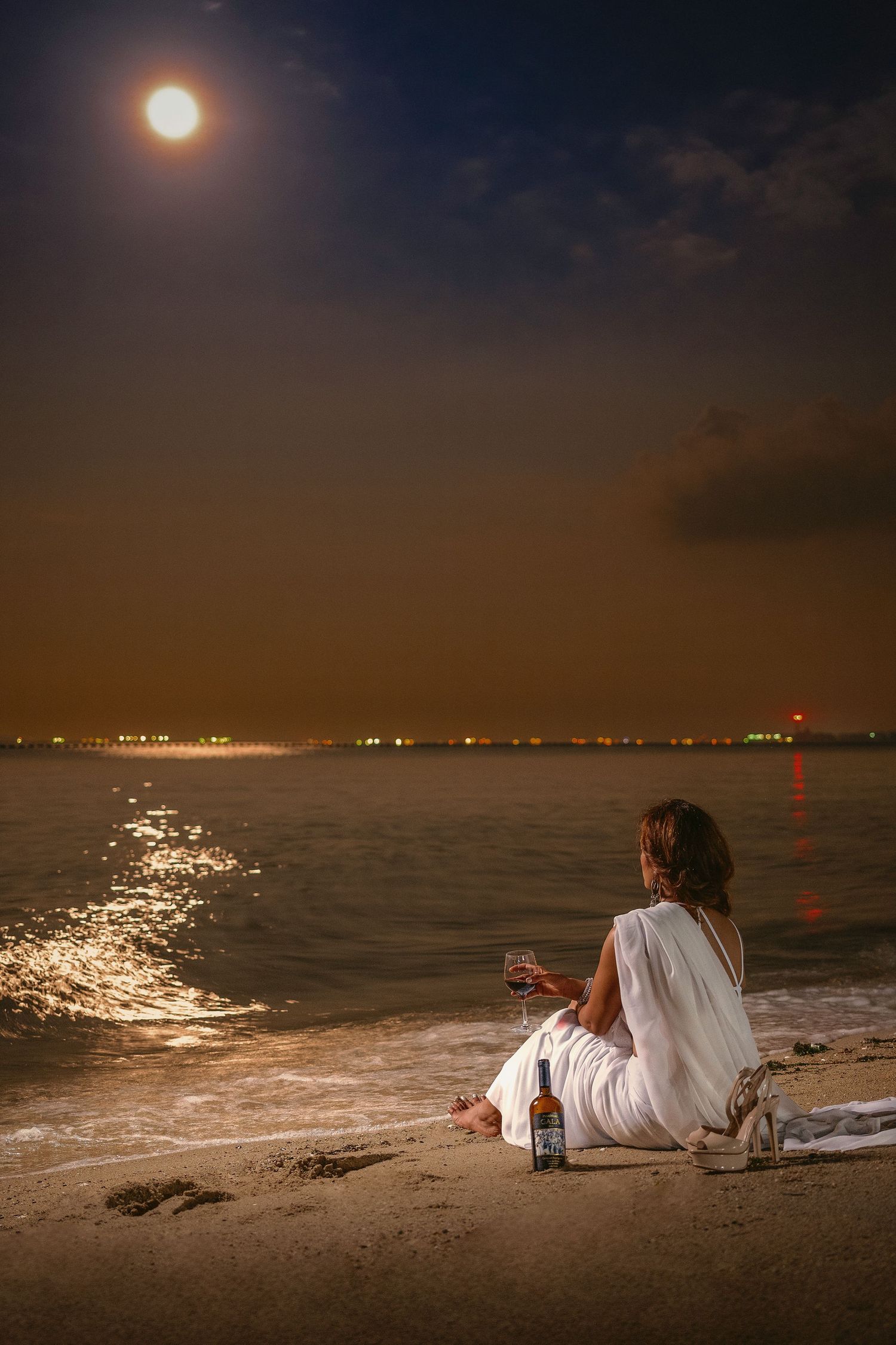 Woman in white dress sitting on moonlit beach at night with wine glass and bottle, city lights reflecting on calm water.