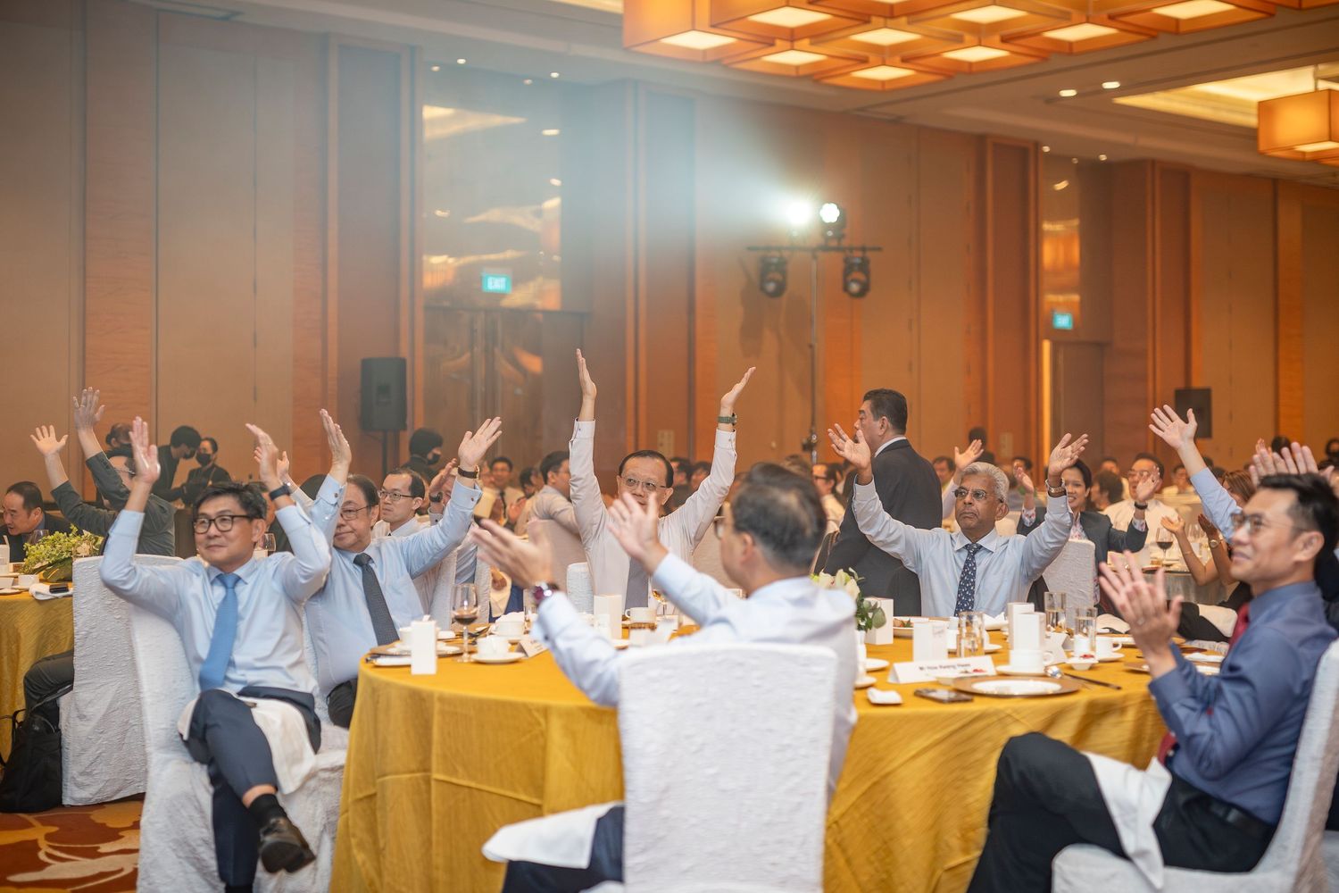 Excited audience members raise hands and applaud at a corporate gala dinner with gold tablecloths.
