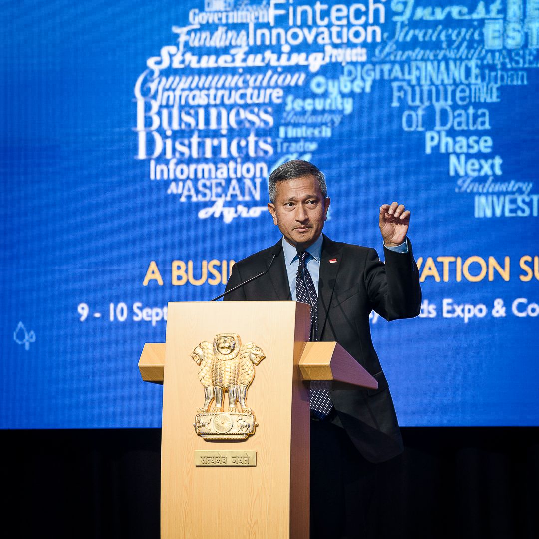 A man in a suit speaks at a podium during a Business Innovation Summit with a digital word cloud backdrop.