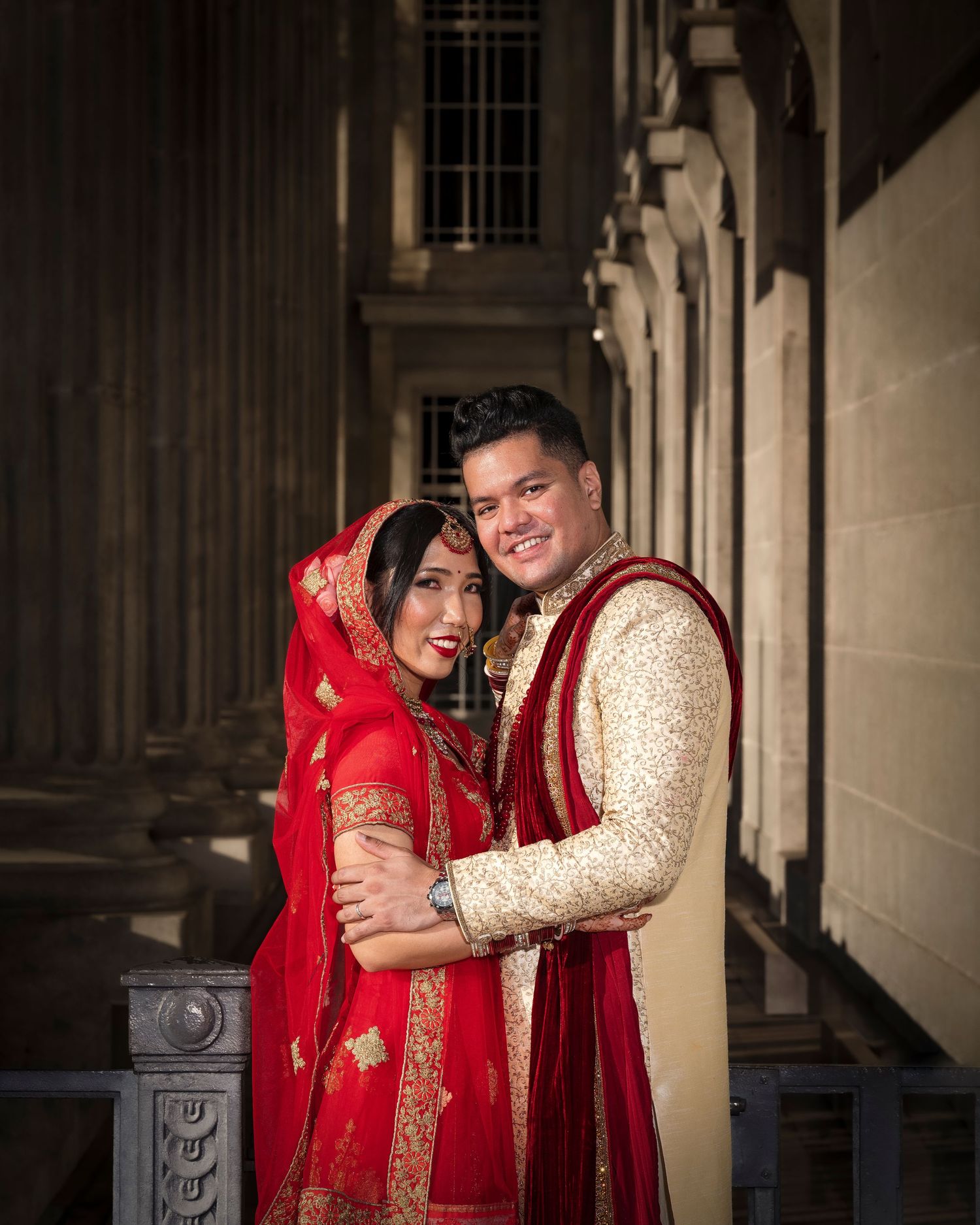 A couple in traditional South Asian wedding attire, bride in red dupatta and groom in cream sherwani, posing at a grand venue.