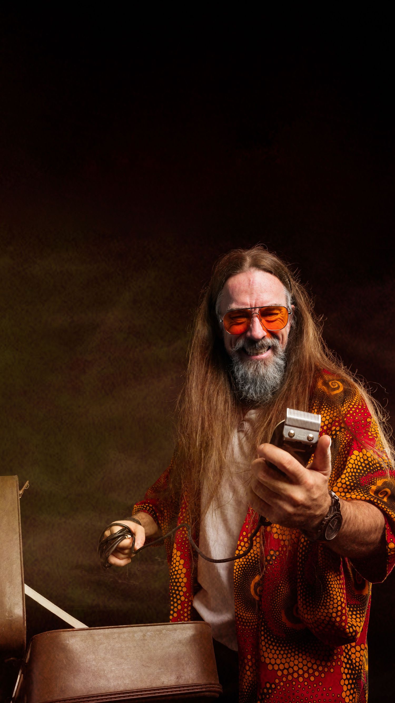 Smiling long-haired man with beard wearing orange sunglasses and vibrant red shirt holding hair clippers.
