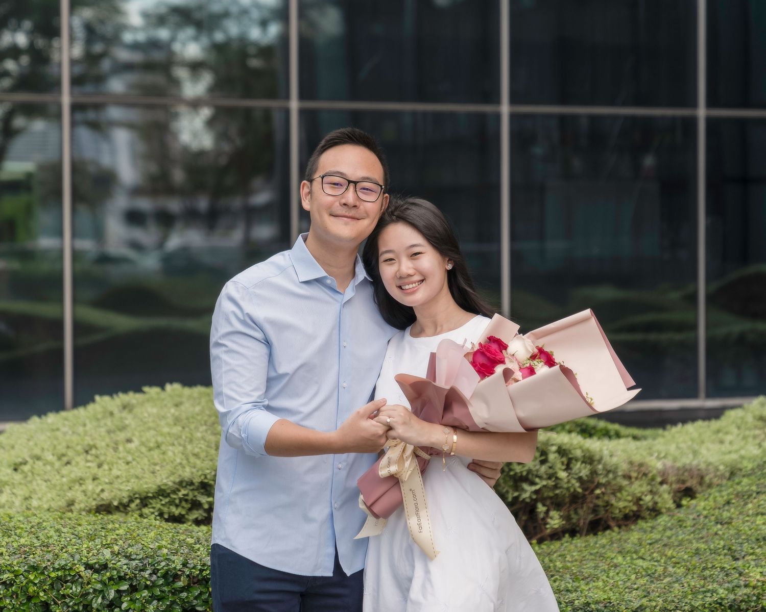 A smiling couple poses outdoors, man embracing woman holding pink flower bouquet, green hedges and glass building behind.