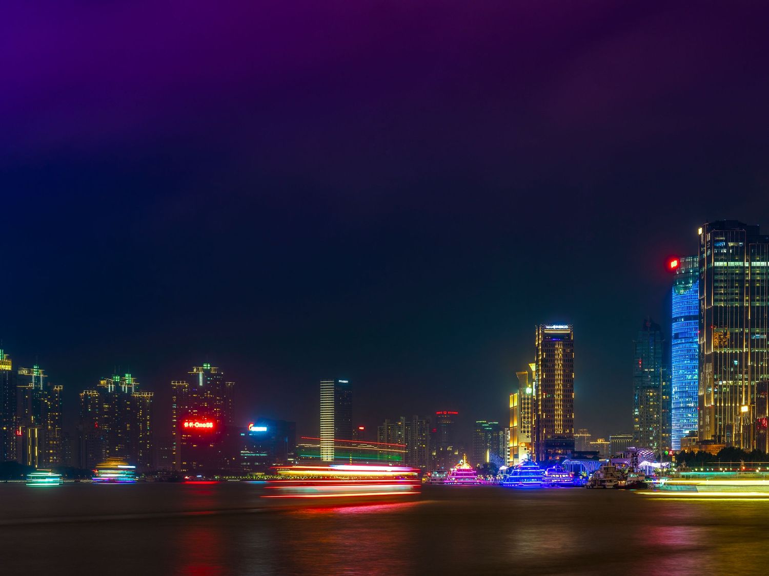 Vibrant city skyline at night with colorful neon lights reflecting on the water and light trails from passing boats.