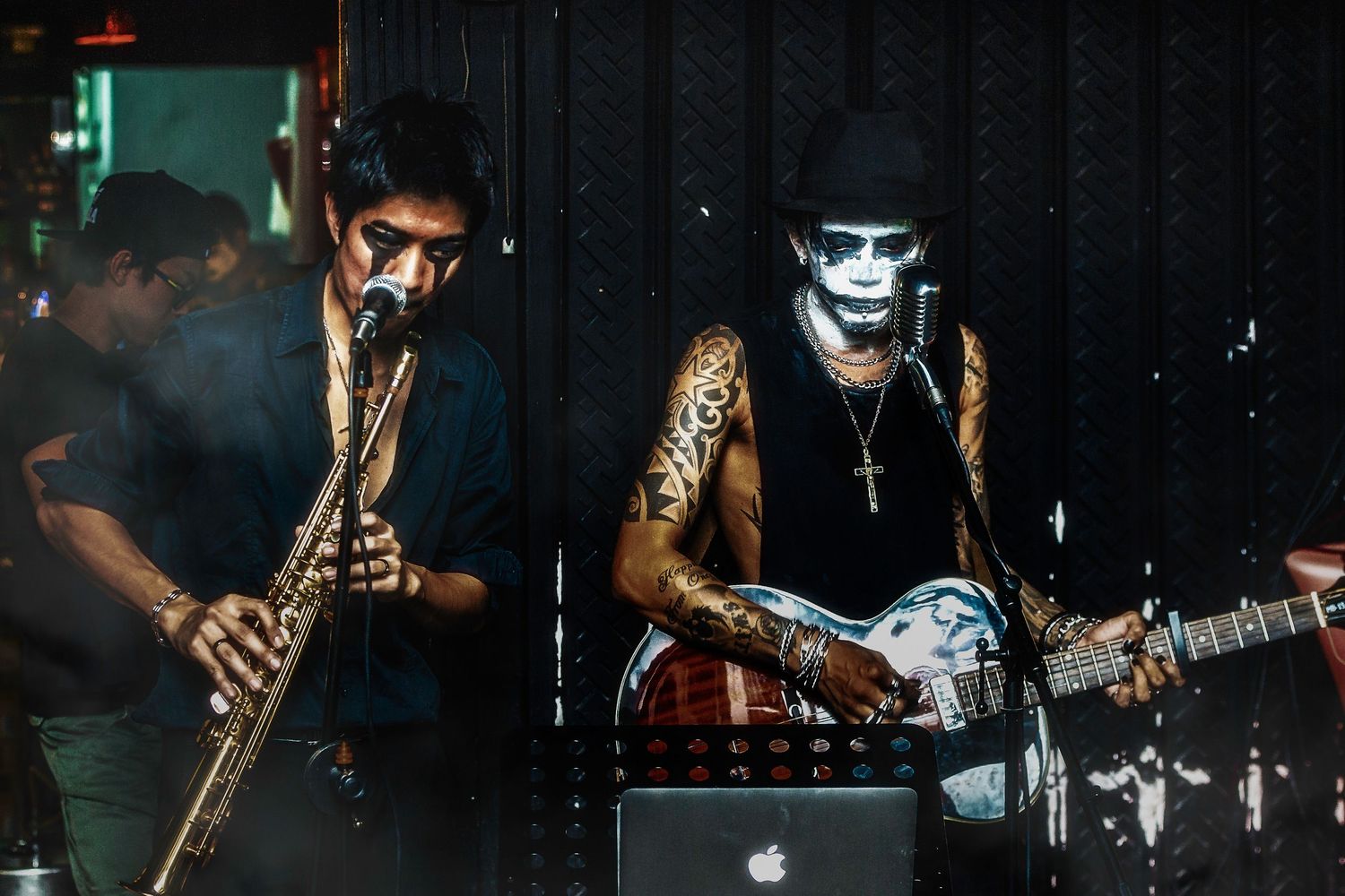 Rock band performing on dark stage with guitarist wearing skull face paint and tattoos.