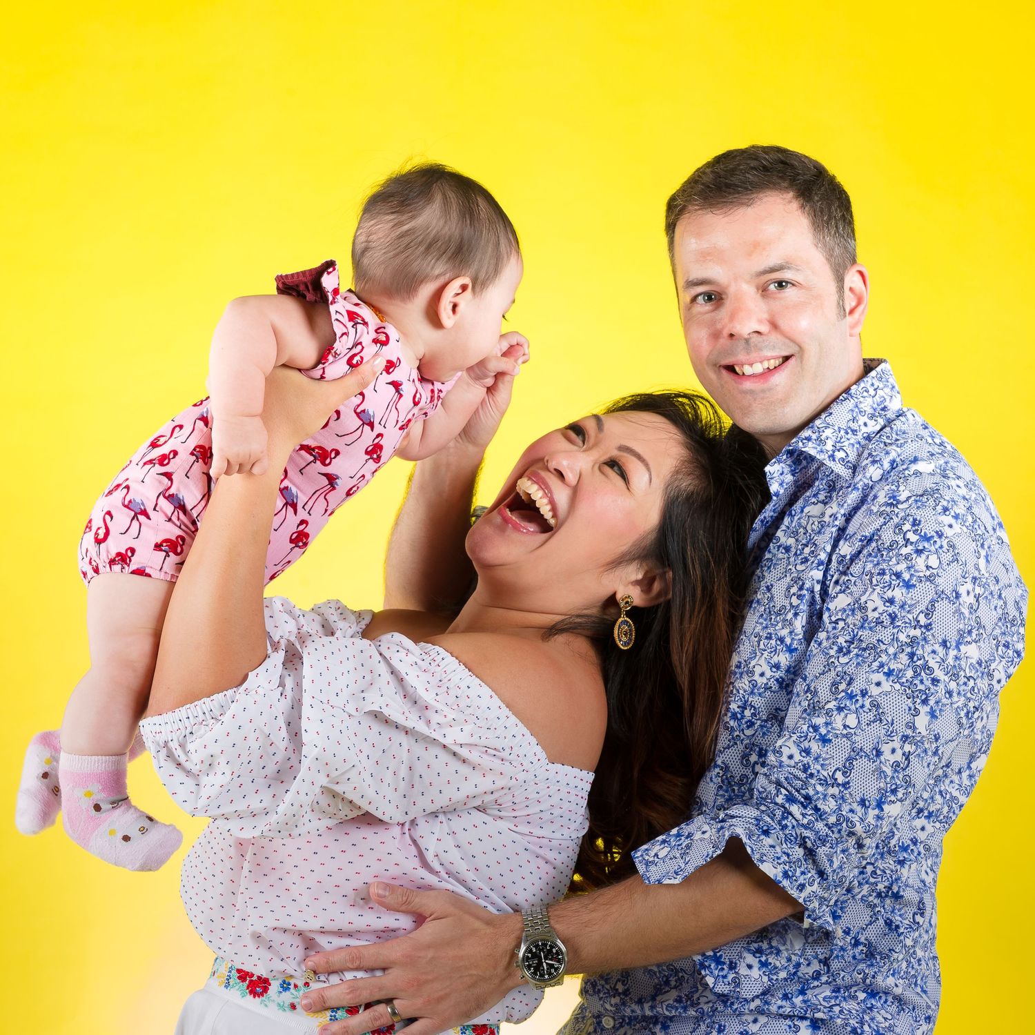 A joyful family poses against a yellow background, with parents laughing as they lift their baby girl in a floral outfit.