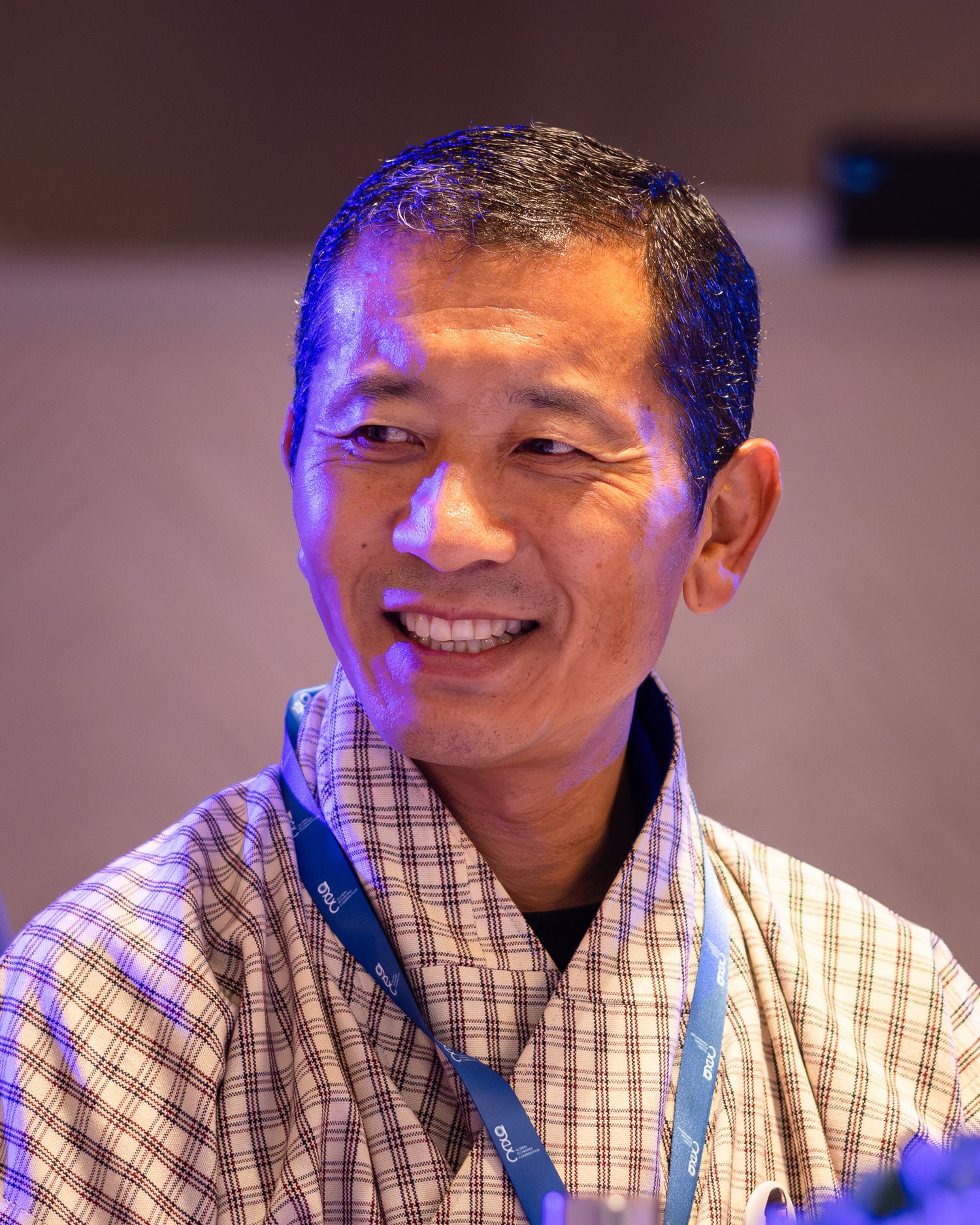Smiling Asian man wearing a plaid shirt and blue lanyard badge at a professional conference event.