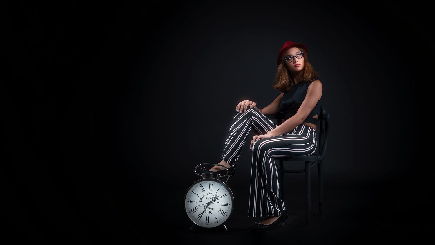 Young woman in striped wide-leg pants and red beret sitting on chair beside large vintage alarm clock.