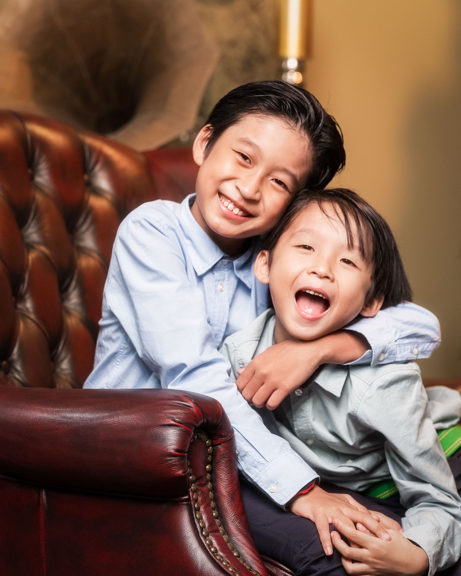 Two young Asian boys laughing and hugging on a leather armchair in a warmly lit room.