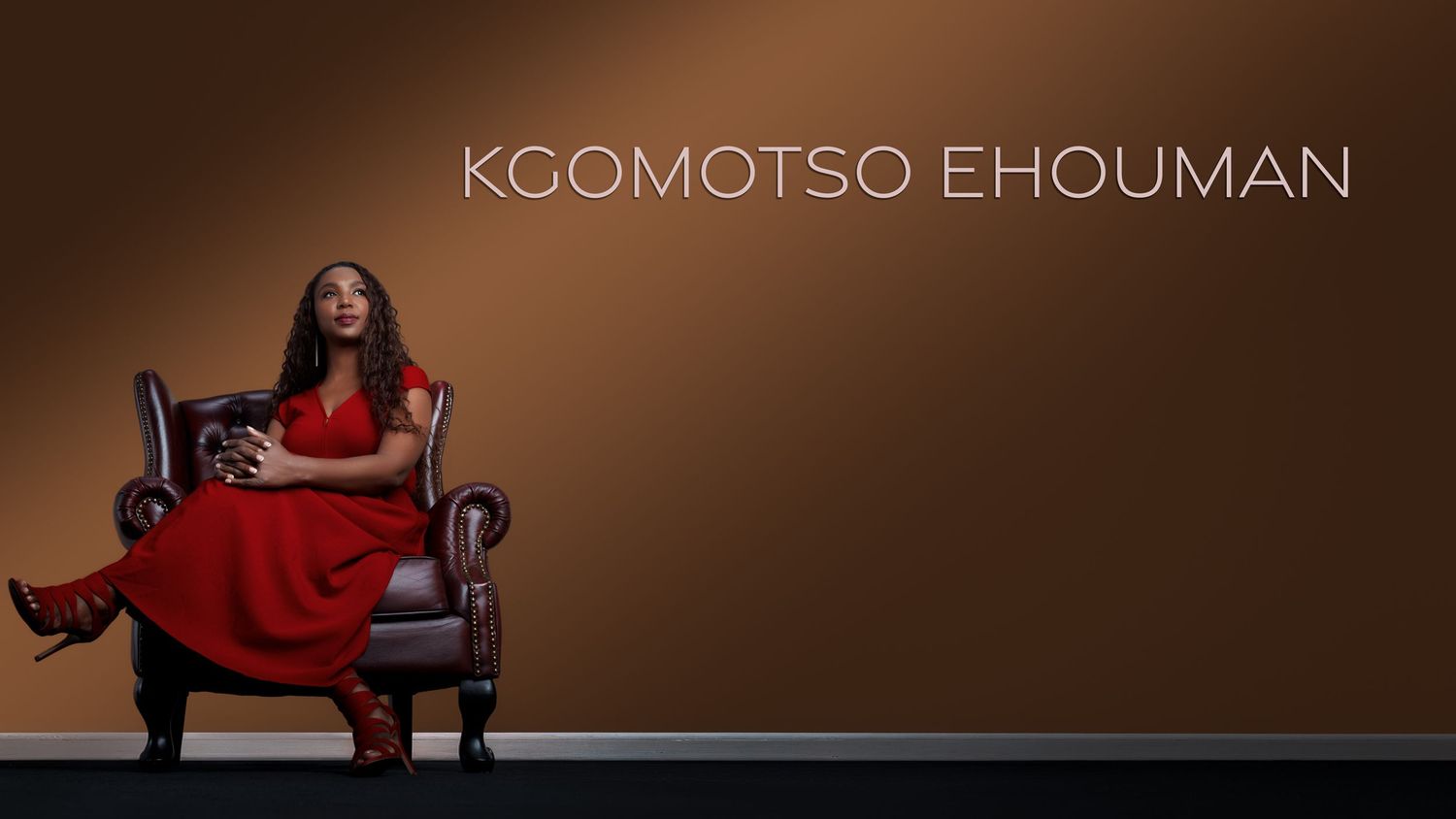 Kgomotso Ehouman sits in a leather chair wearing a red dress and boots against a brown studio background.