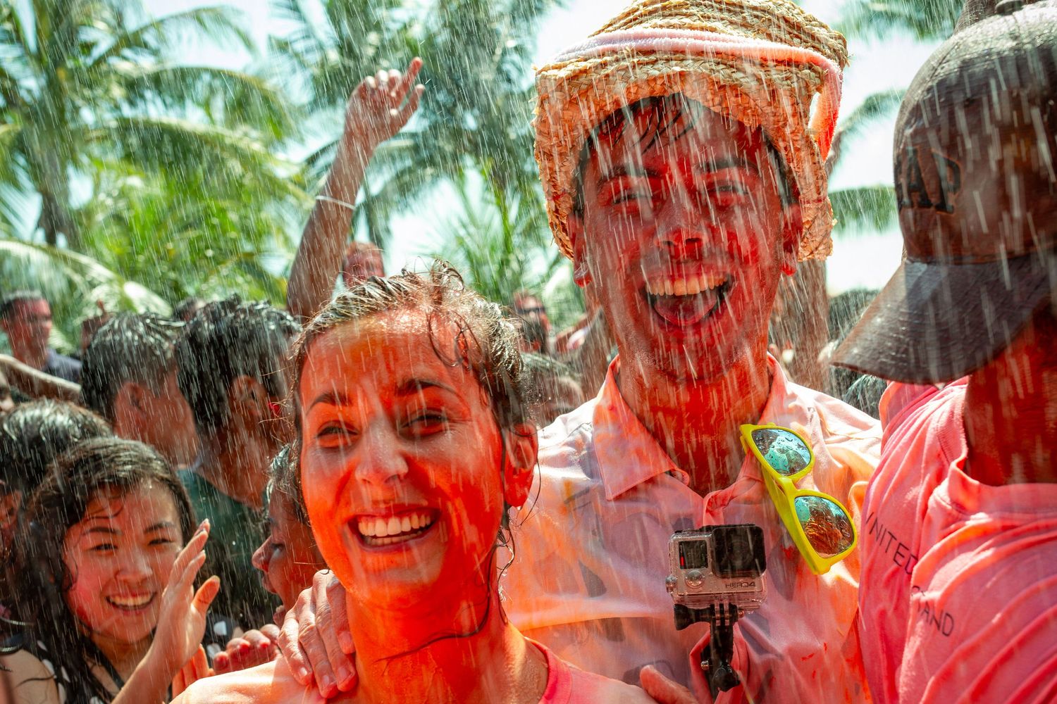 People covered in colorful paint laugh and celebrate outdoors during a festive Holi-style color festival near palm trees.
