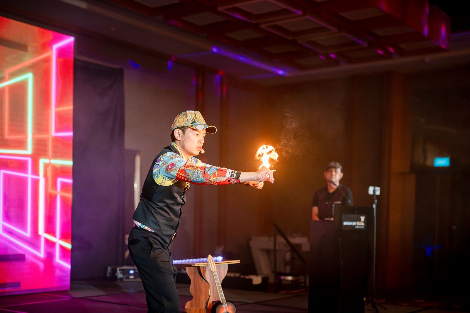 Tattooed fire performer holds flames during a colorful neon-lit stage show at an indoor event.