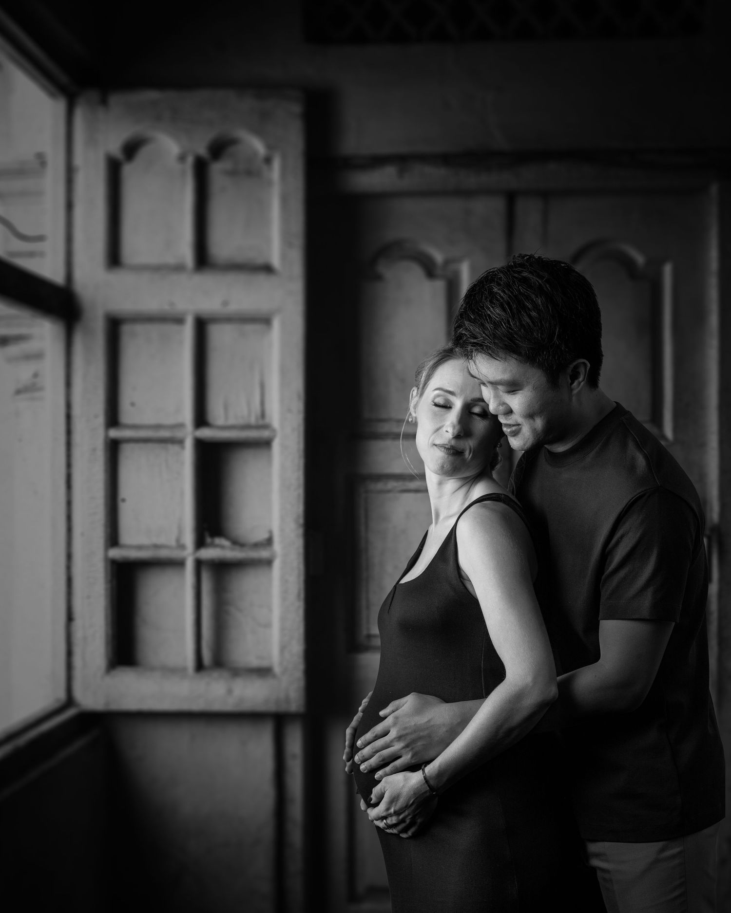 Black and white photo of a pregnant couple embracing tenderly near a vintage window in a dimly lit room.