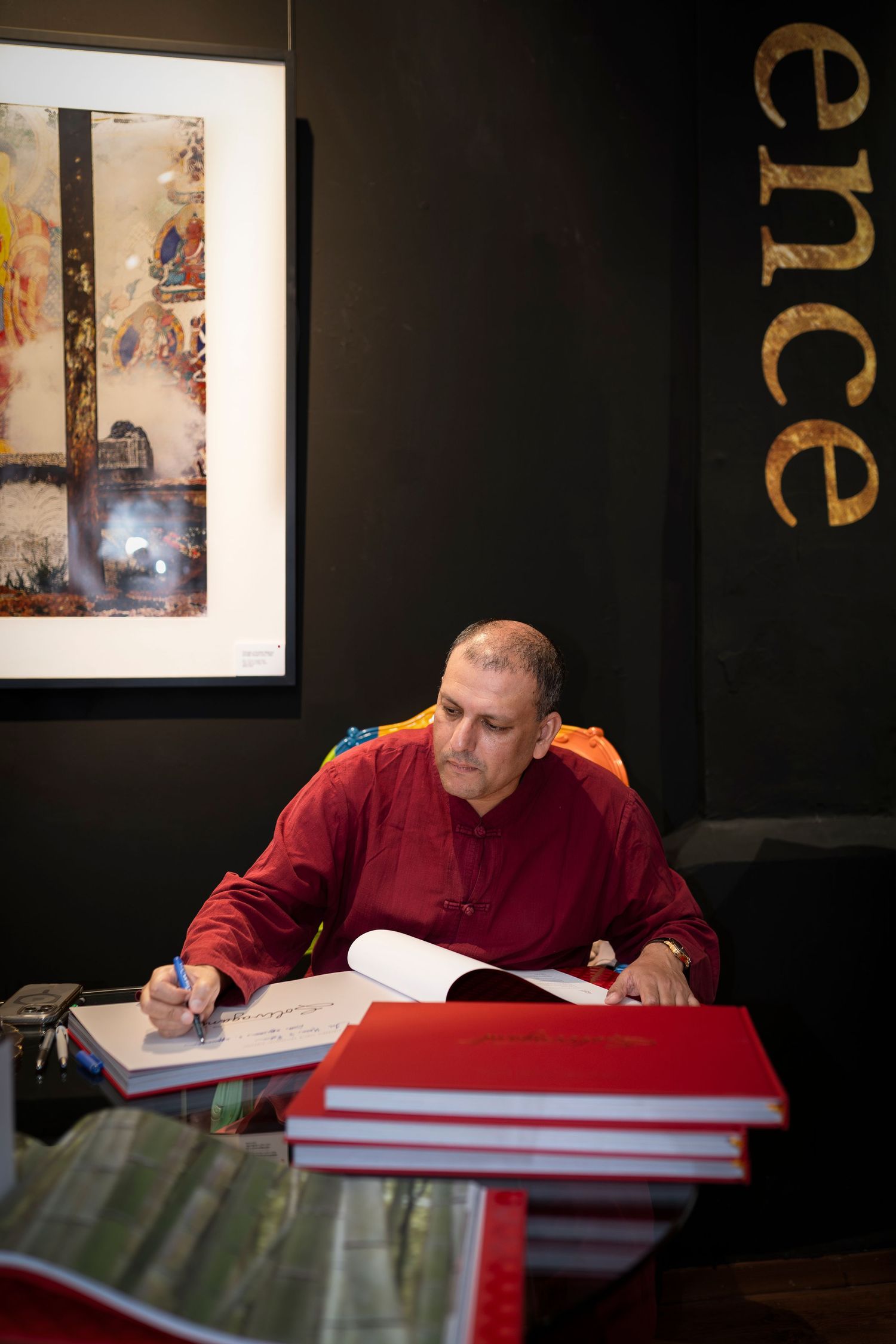 A bald man in a red shirt writes in a large book at a desk, surrounded by artwork in a dark gallery.