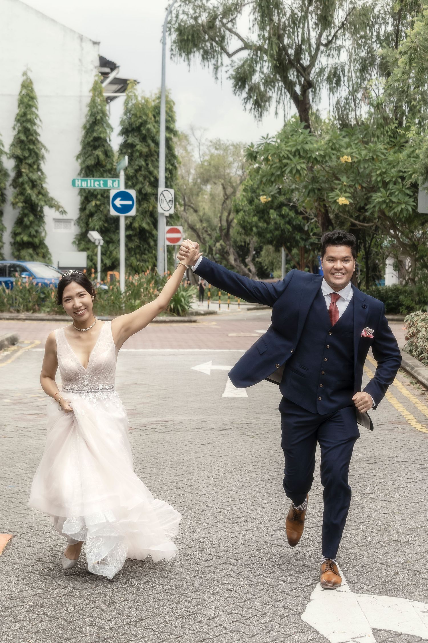 Joyful newlywed couple in wedding attire running hand-in-hand on a street, bride in white gown, groom in navy suit.