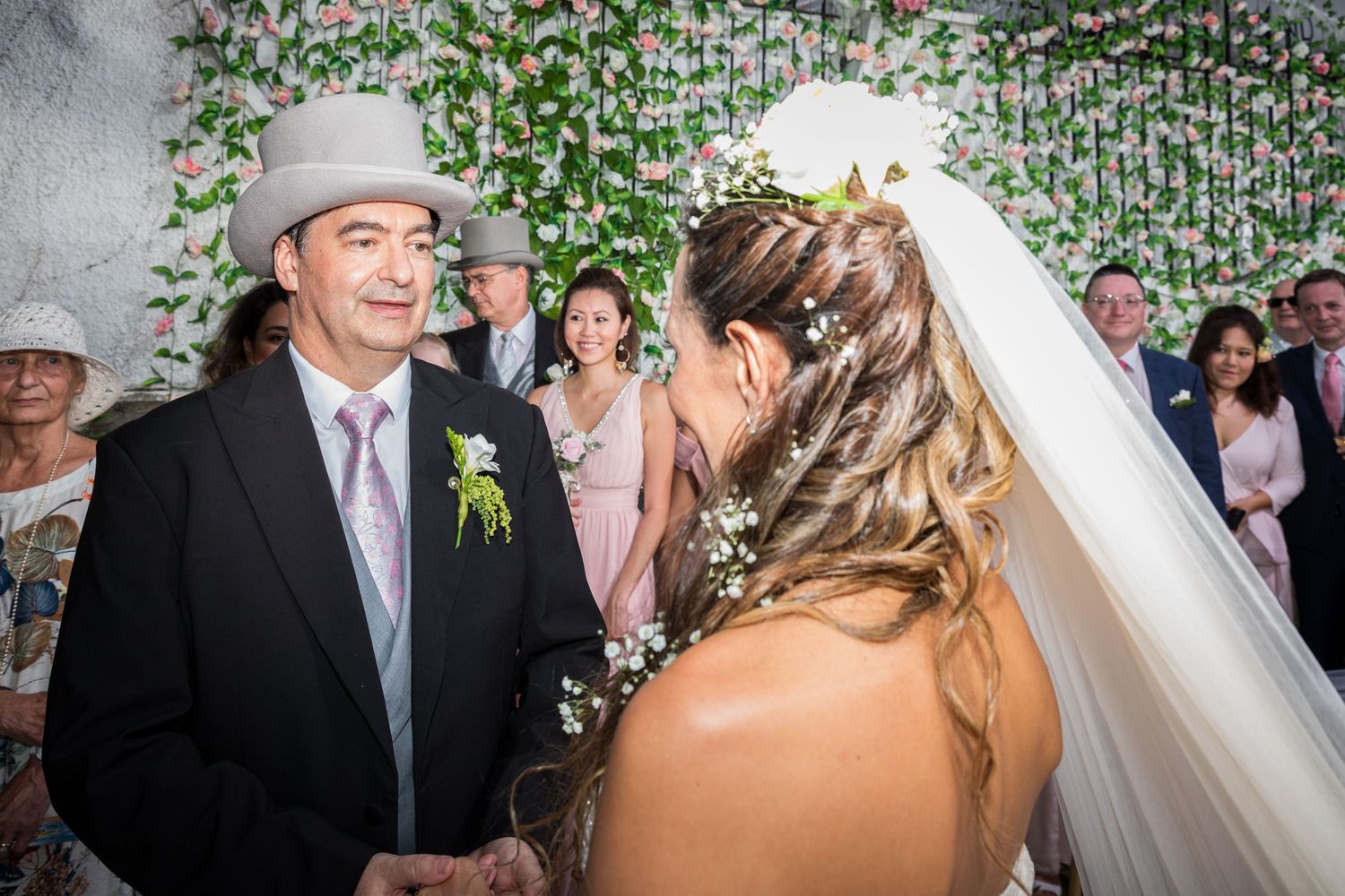 Man in cowboy hat smiling at bride in veil at wedding ceremony with floral wall backdrop.
