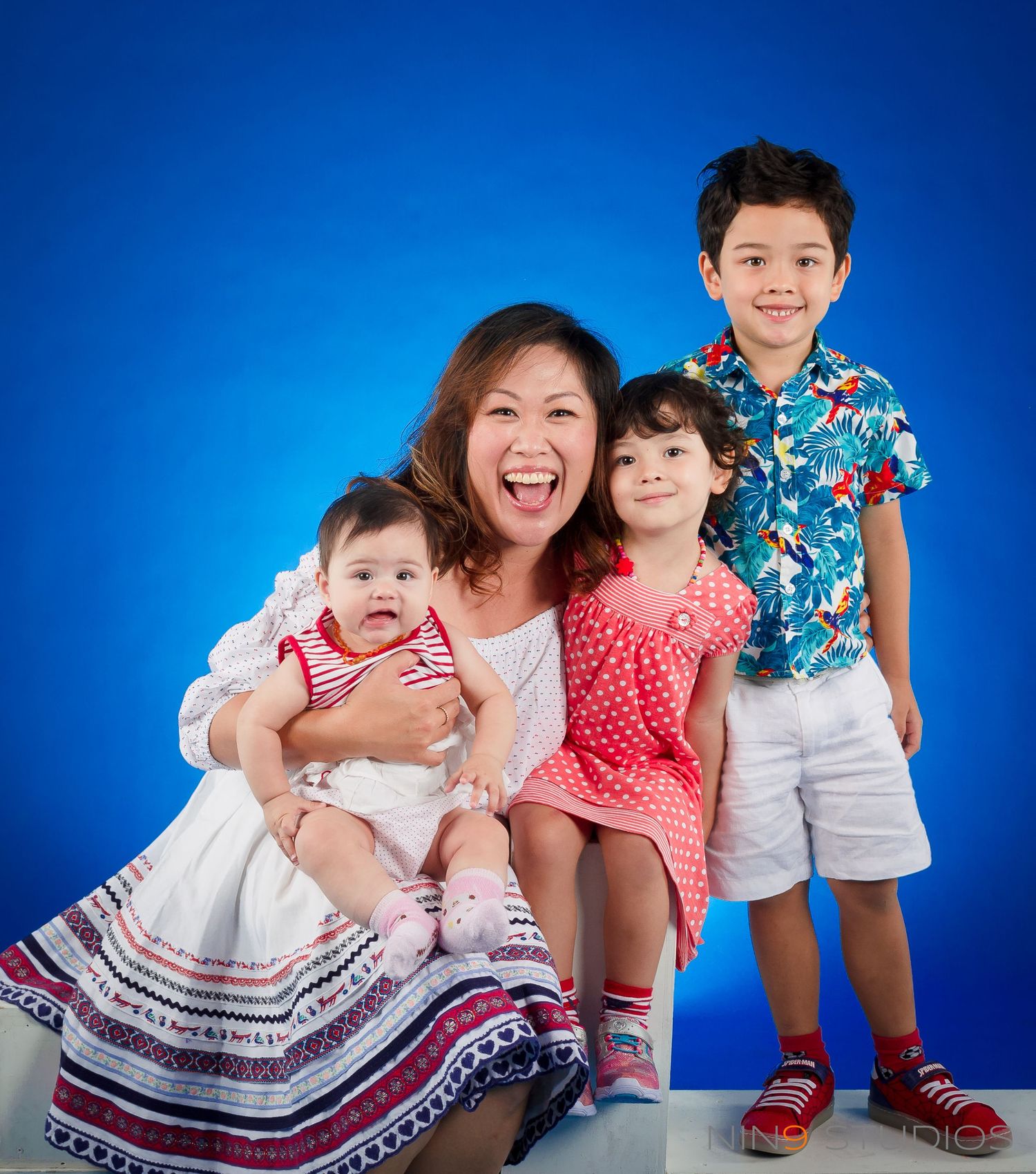 A smiling Asian mother poses with her three young children against a bright blue background in a cheerful family portrait.