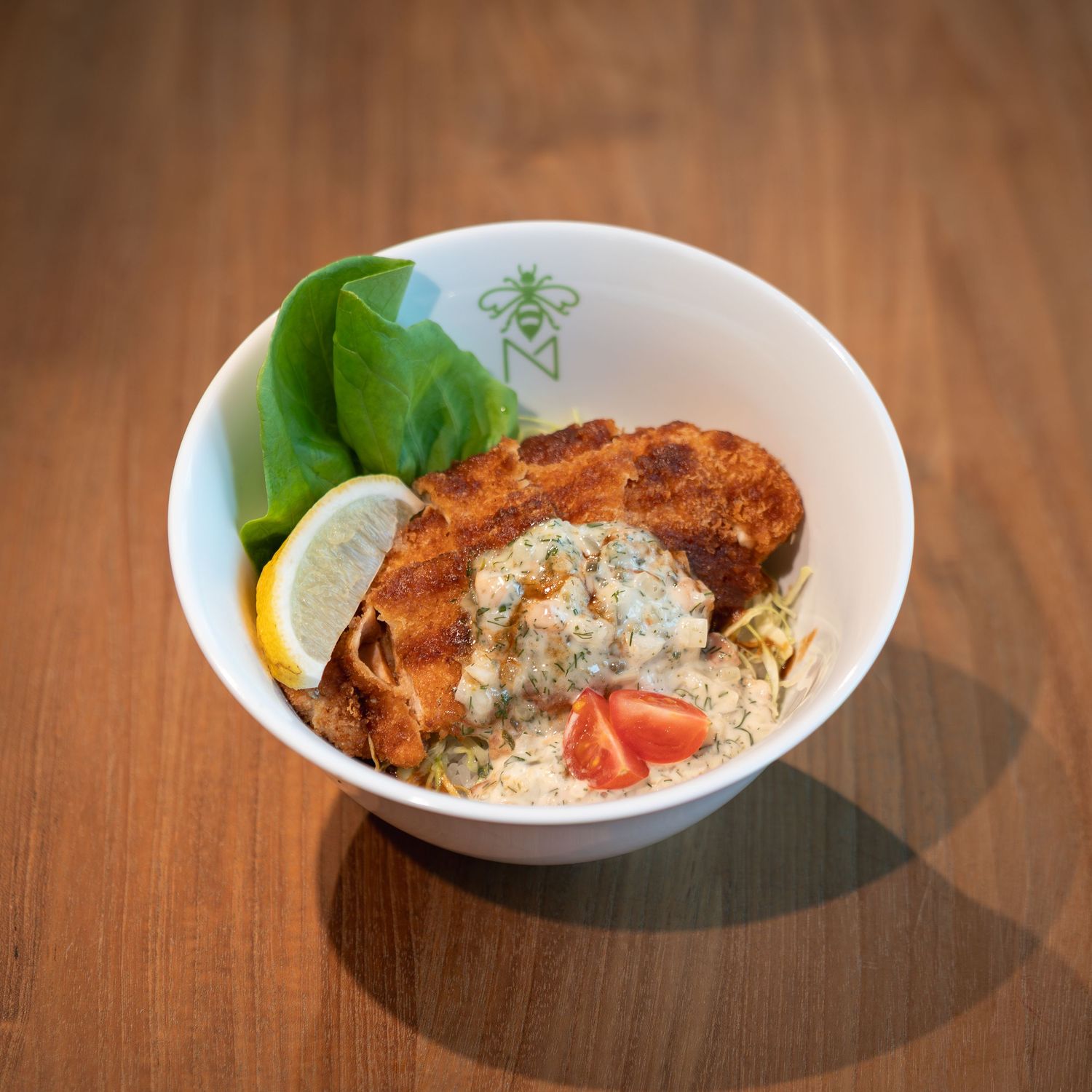 Crispy breaded chicken in a white bowl with creamy herb sauce, fresh tomato, lemon slice, and basil leaves.