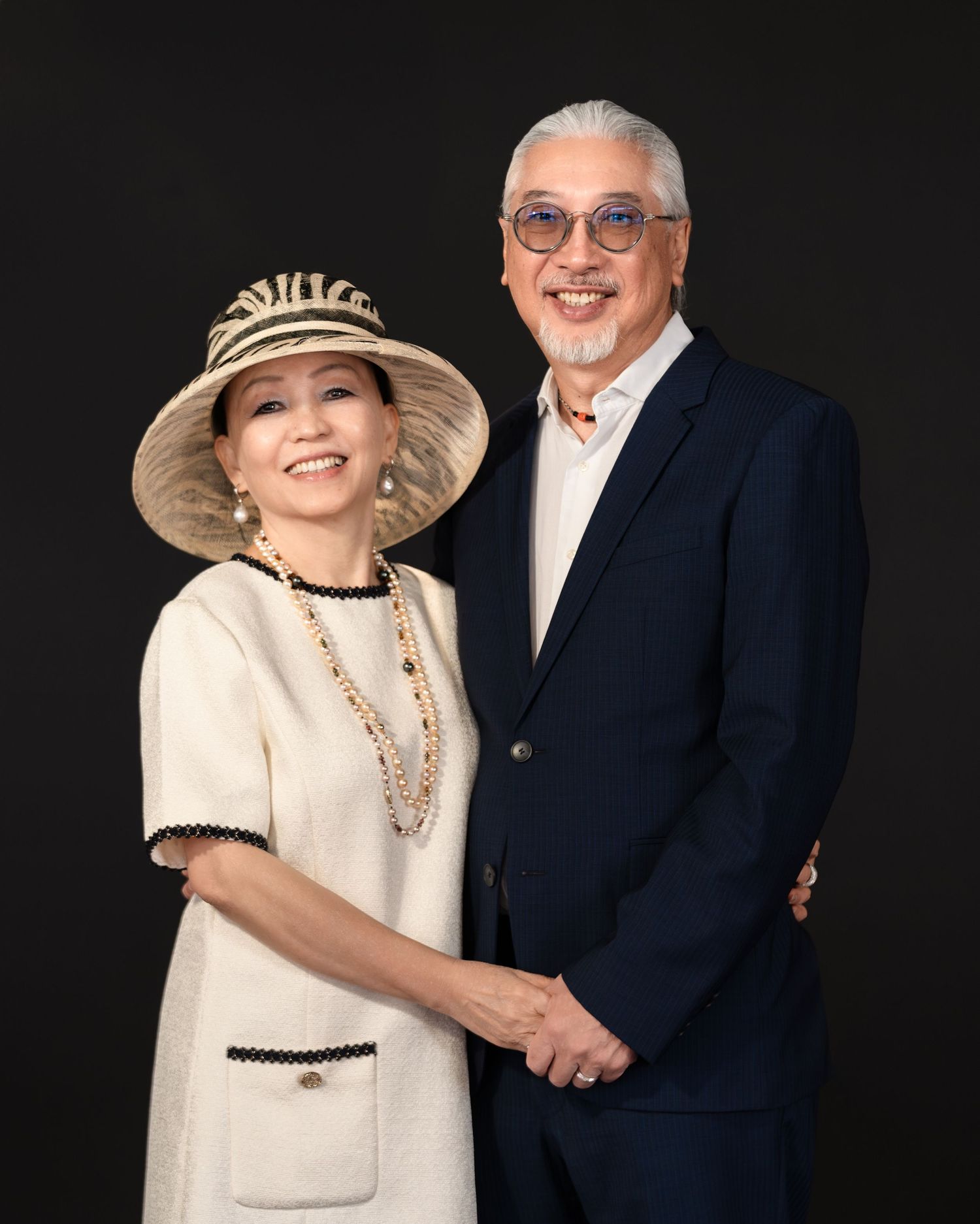 Elegant couple posing together, woman in cream dress and wide-brim hat, man in navy suit, against dark background.