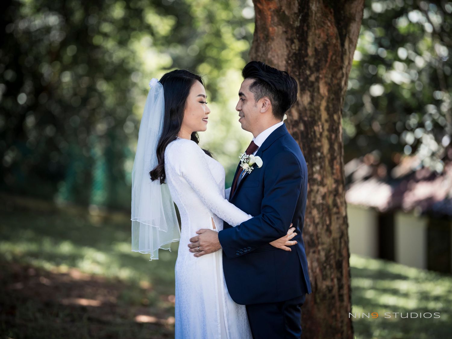 A bride in a white ao dai and groom in navy suit share a romantic moment beneath a tree outdoors.