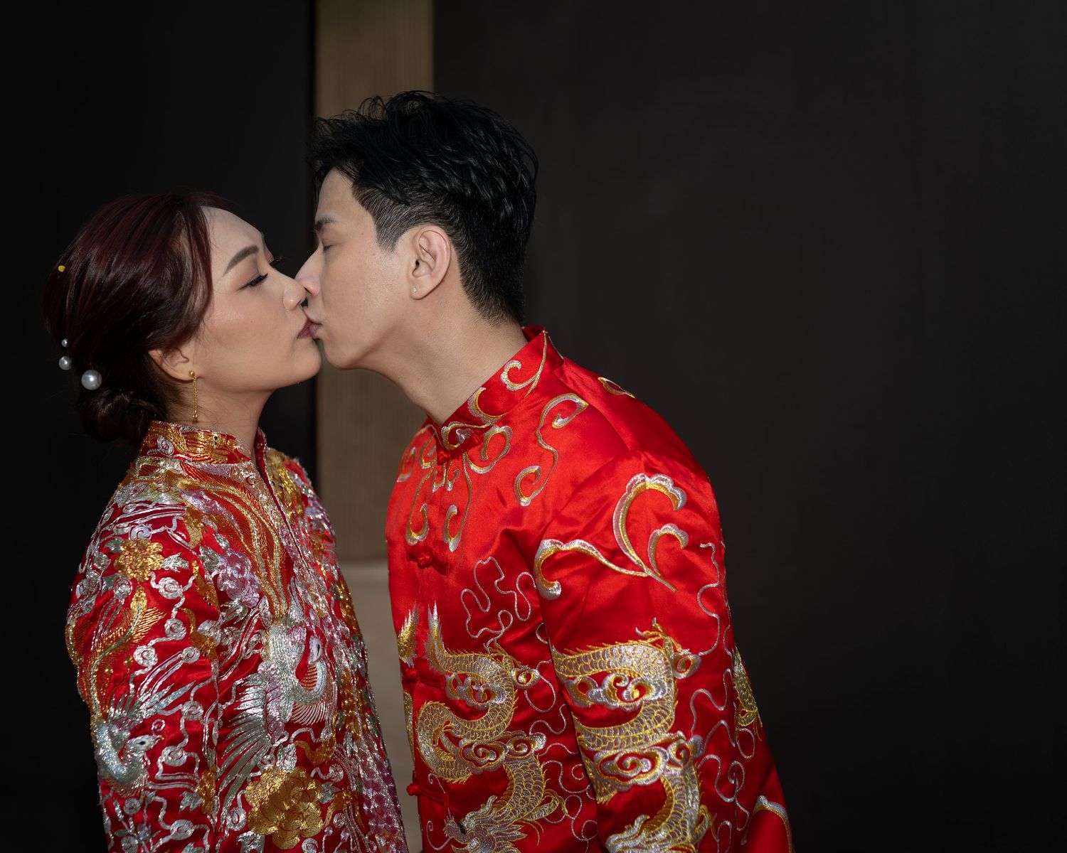 Couple kissing in traditional red Chinese wedding attire with gold dragon embroidery during a romantic ceremony.