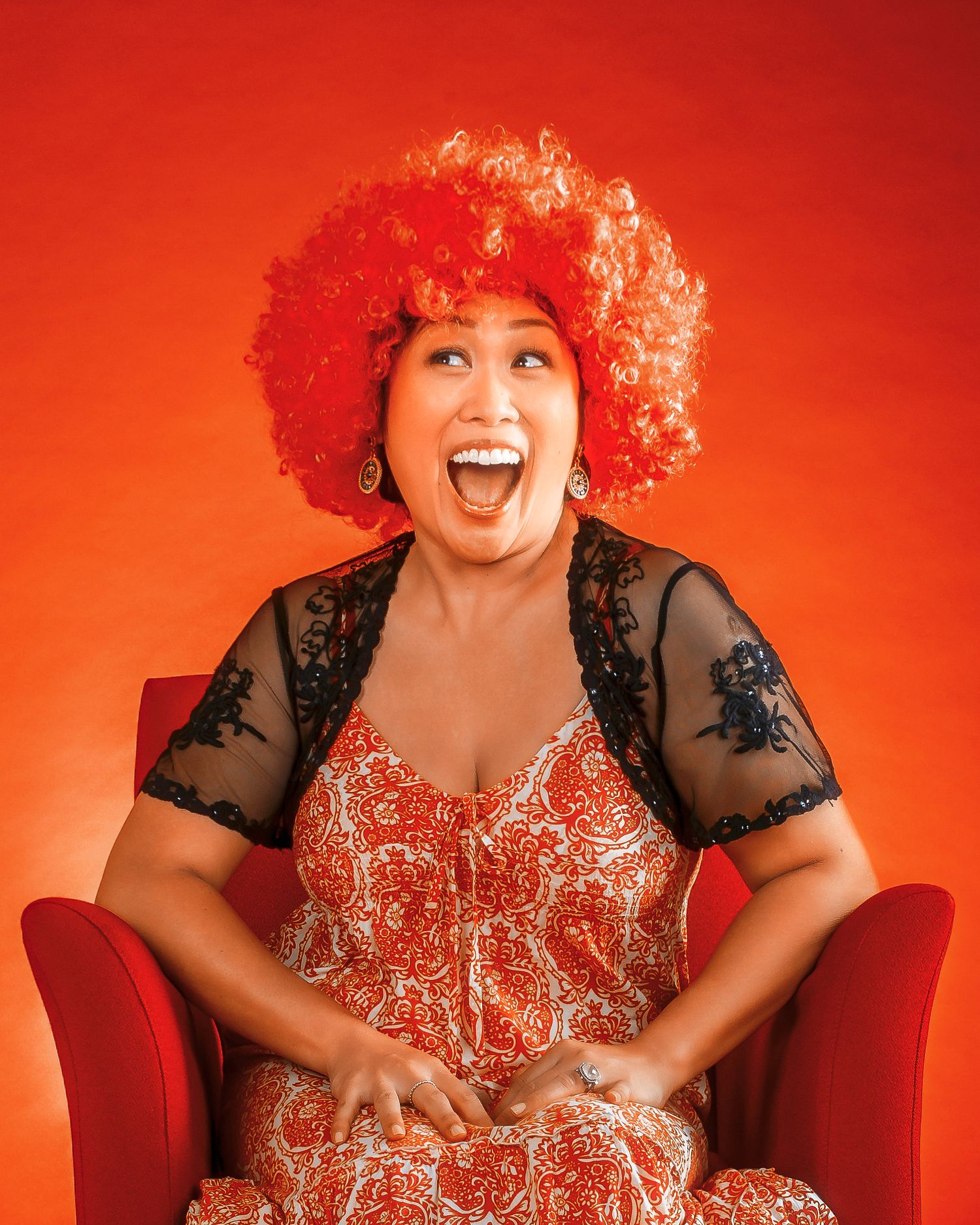 Woman in orange afro wig and floral dress laughing joyfully while seated in red chair against orange background.