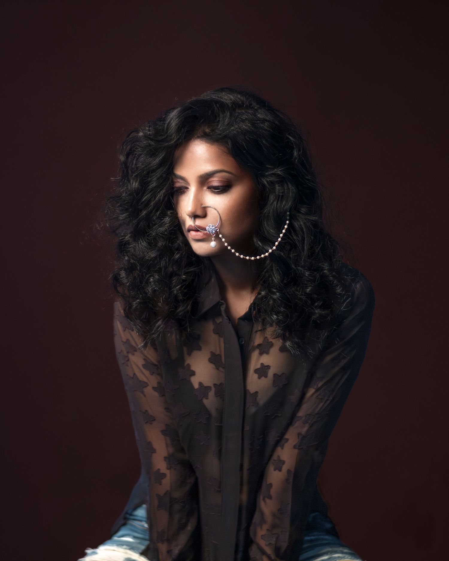 Woman with curly black hair wearing a sheer lace top and large hoop earrings poses against a dark background.