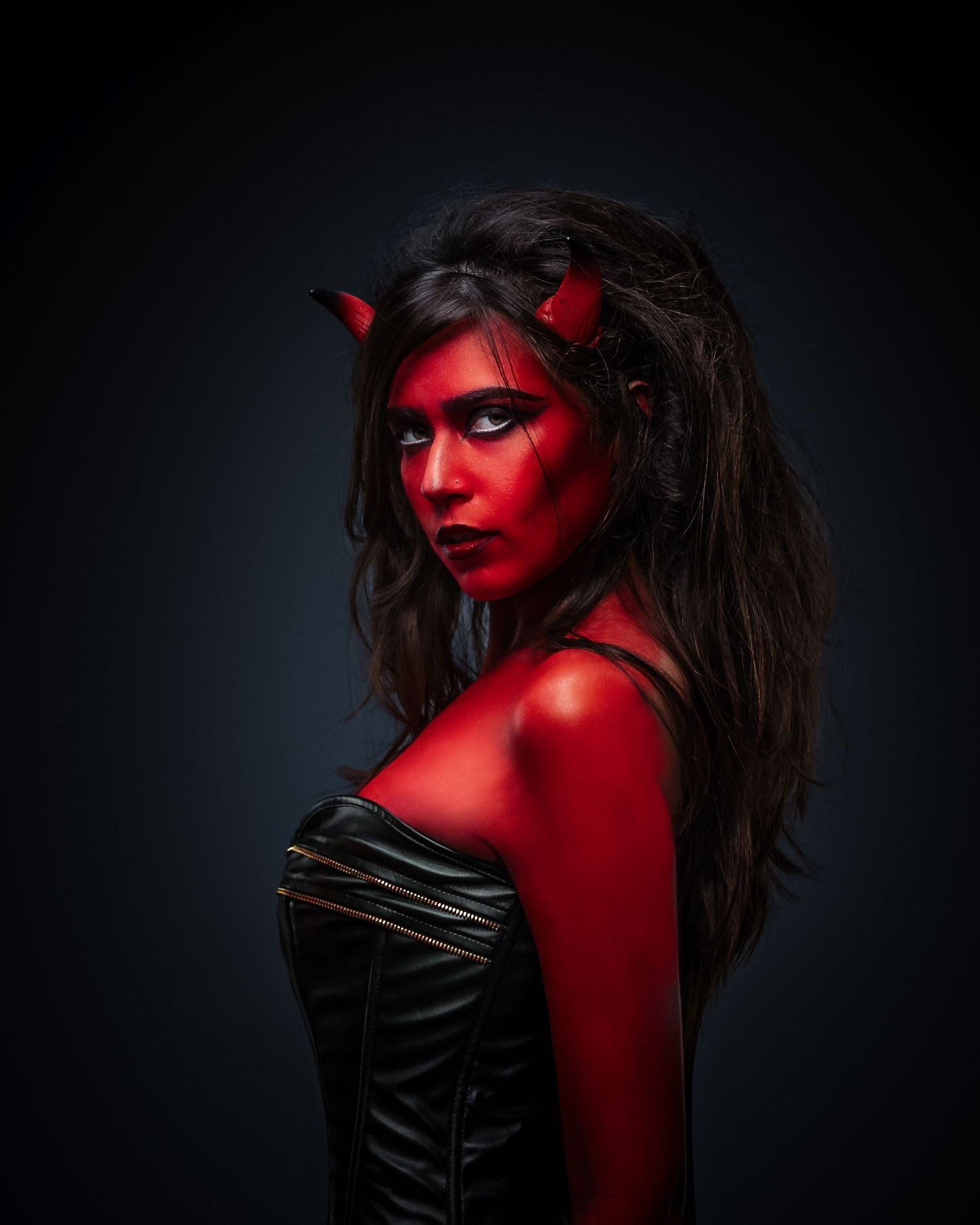 Woman in red body paint and devil horns costume wearing a black leather corset against dark background.