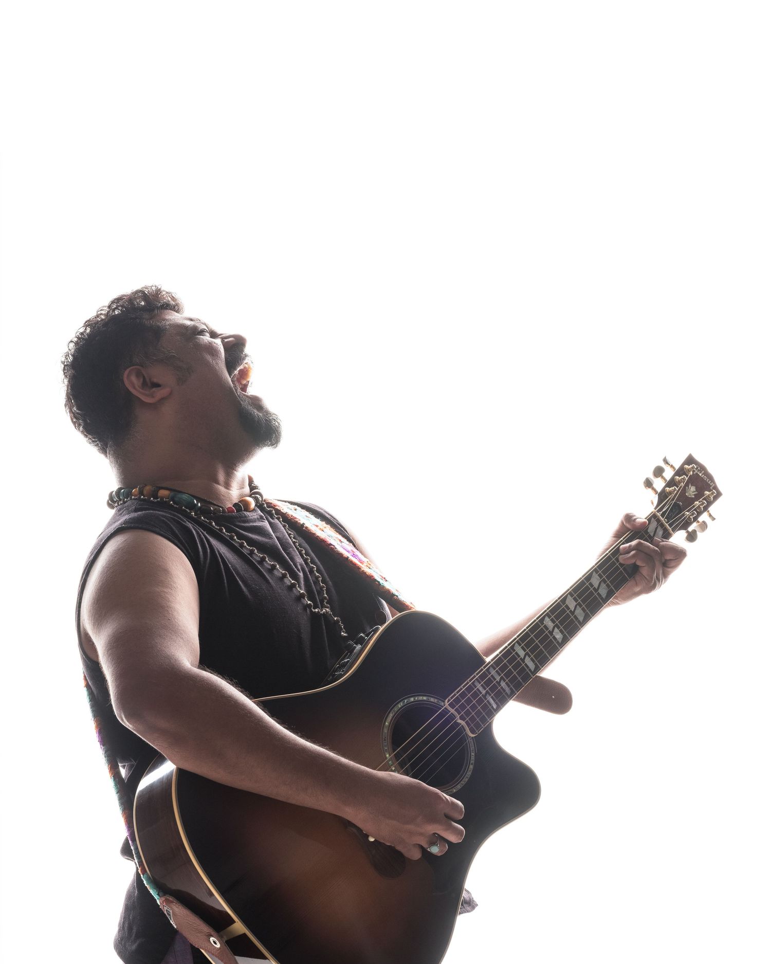 Musician playing acoustic guitar with head tilted back, singing passionately against a bright white background.
