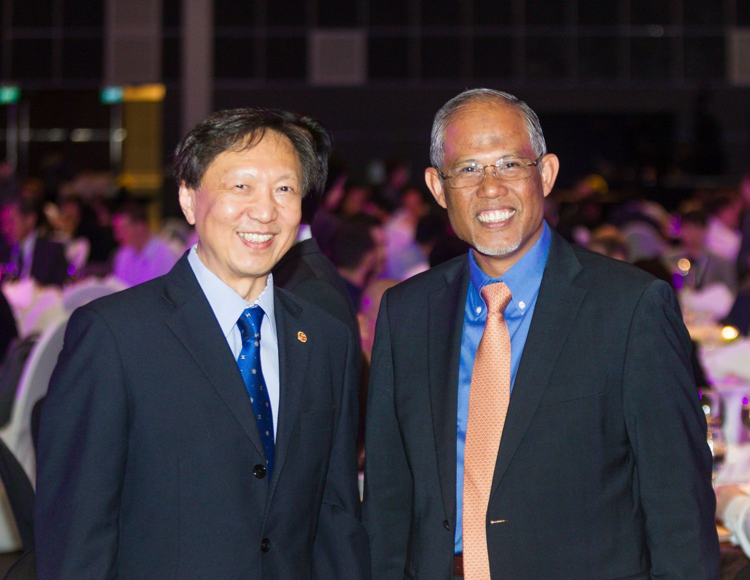 Two men in suits smile together at an elegant formal dinner event with purple lighting.