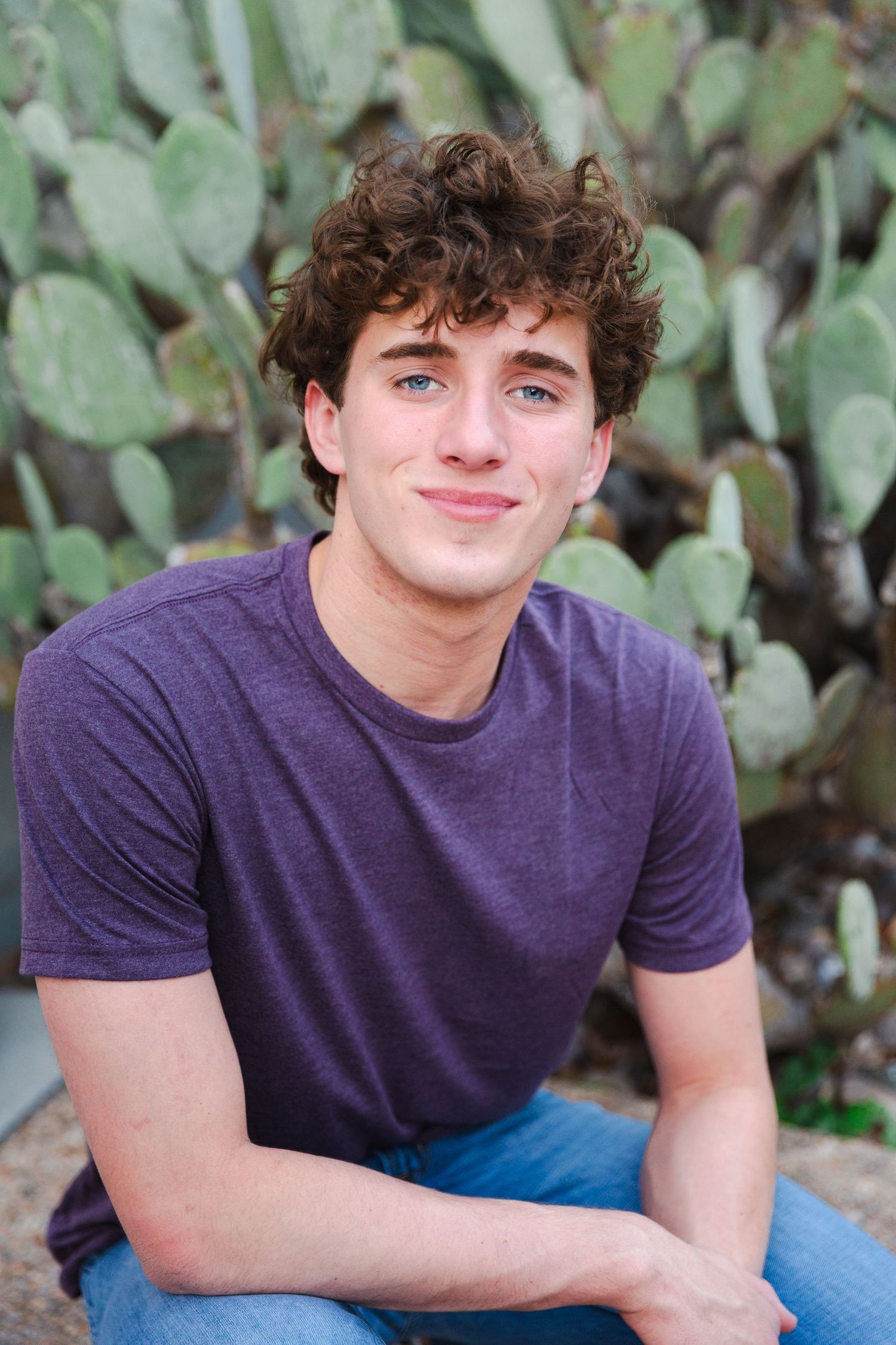 Young man in purple t-shirt smiles while sitting in front of cactus garden.