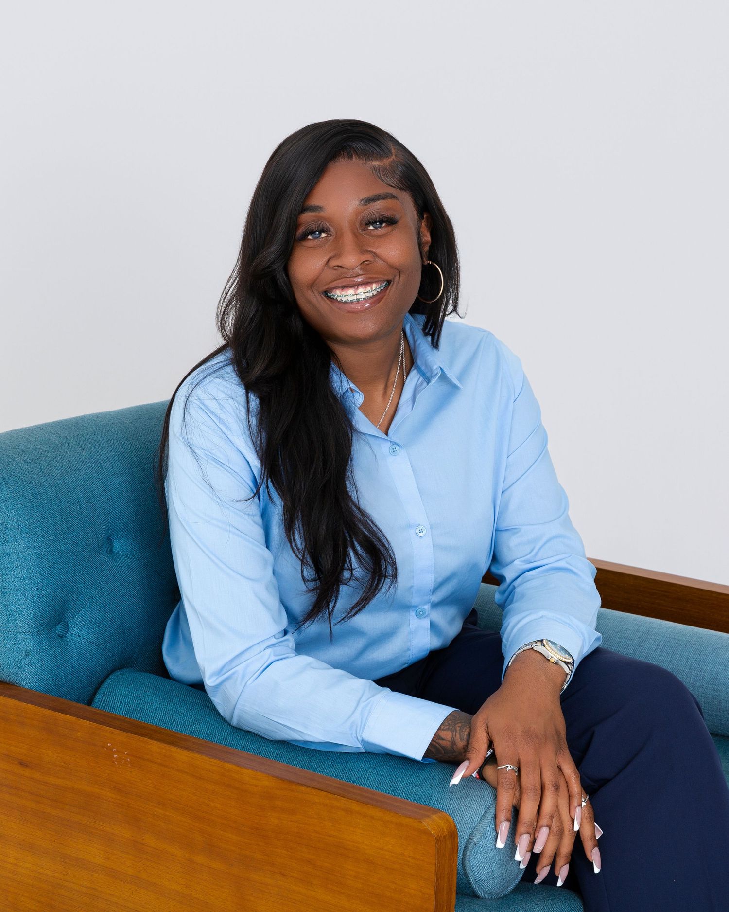 Professional woman in light blue button-down shirt sits on turquoise chair smiling at camera.