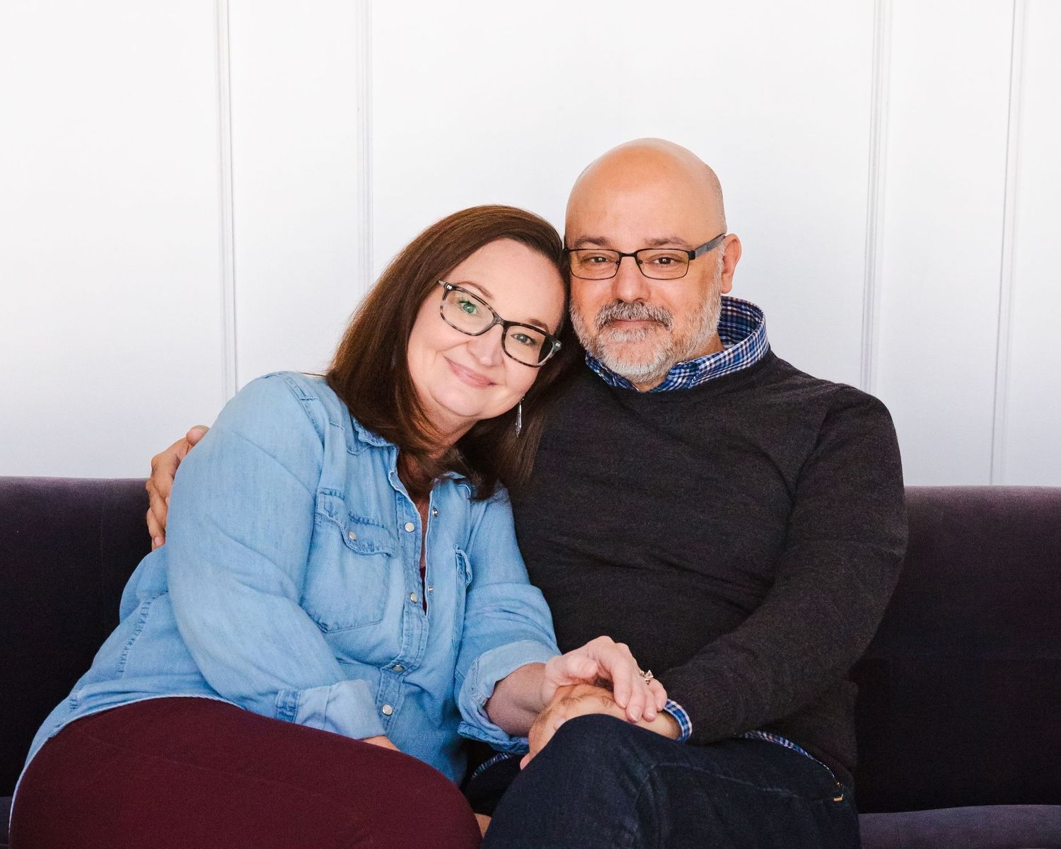 A couple relaxes on a purple couch, dressed casually in denim and dark sweaters.