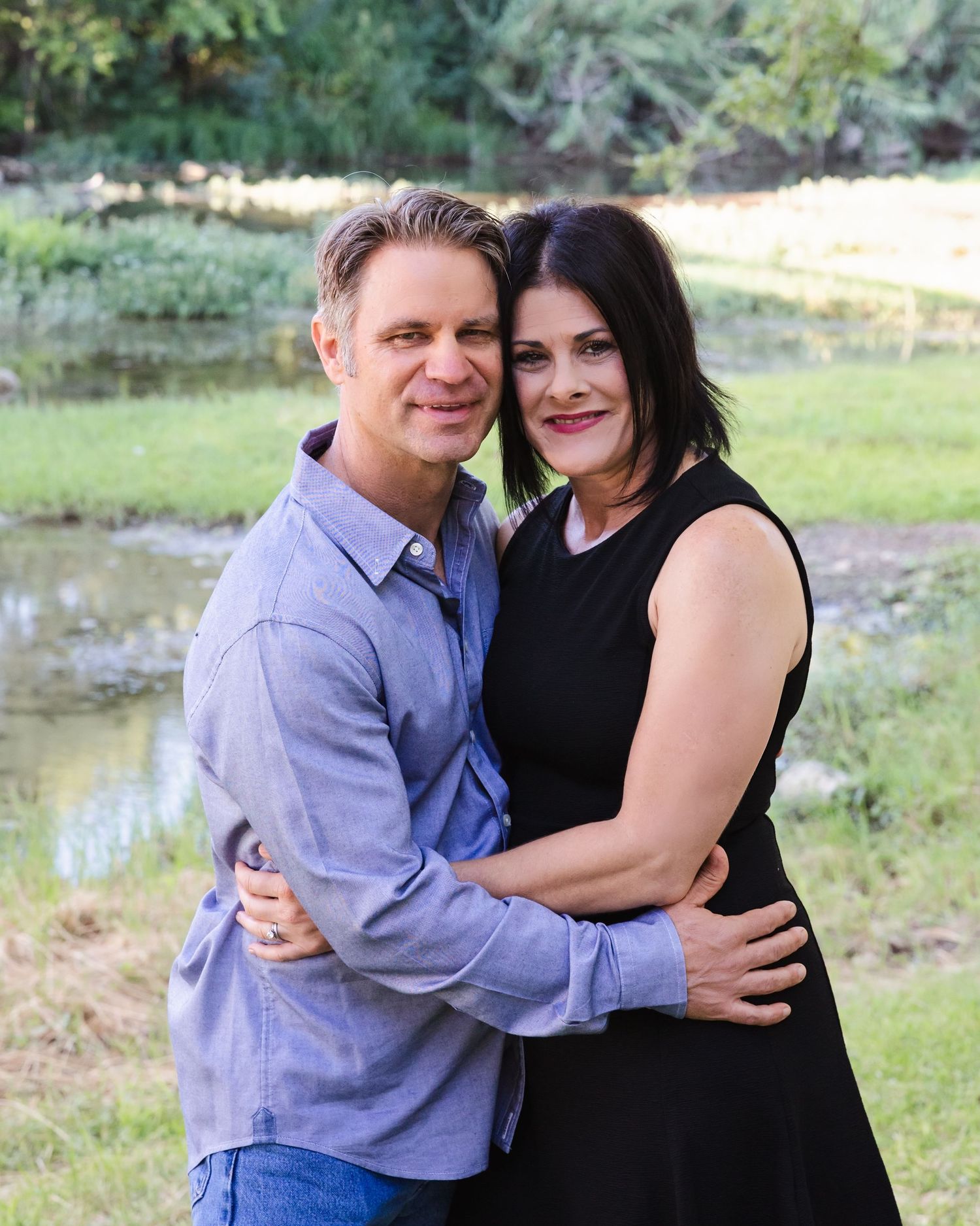 A couple embraces outdoors near a pond, dressed in casual attire against a natural backdrop.
