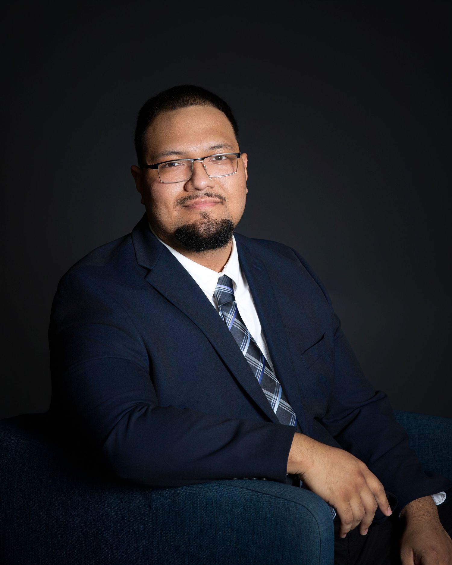 Professional portrait of businessman in navy suit seated against dark background.