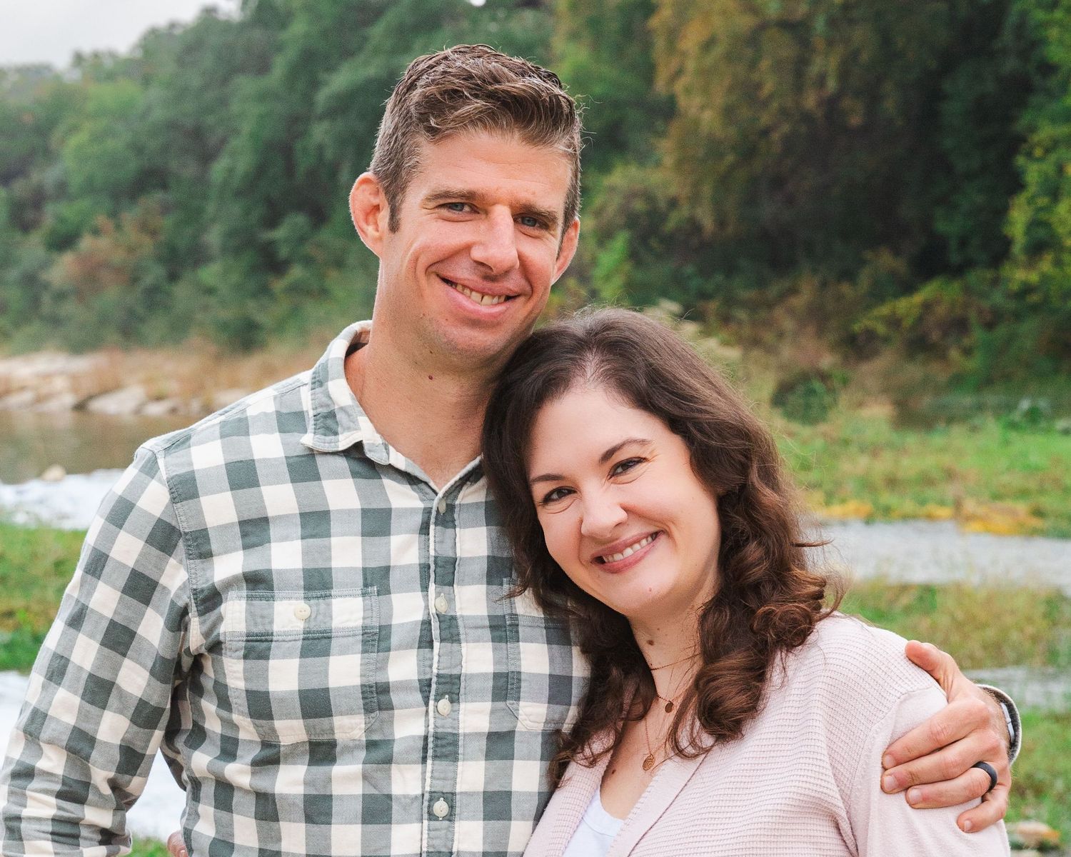 A couple poses together outdoors in casual attire with autumn foliage in the background.