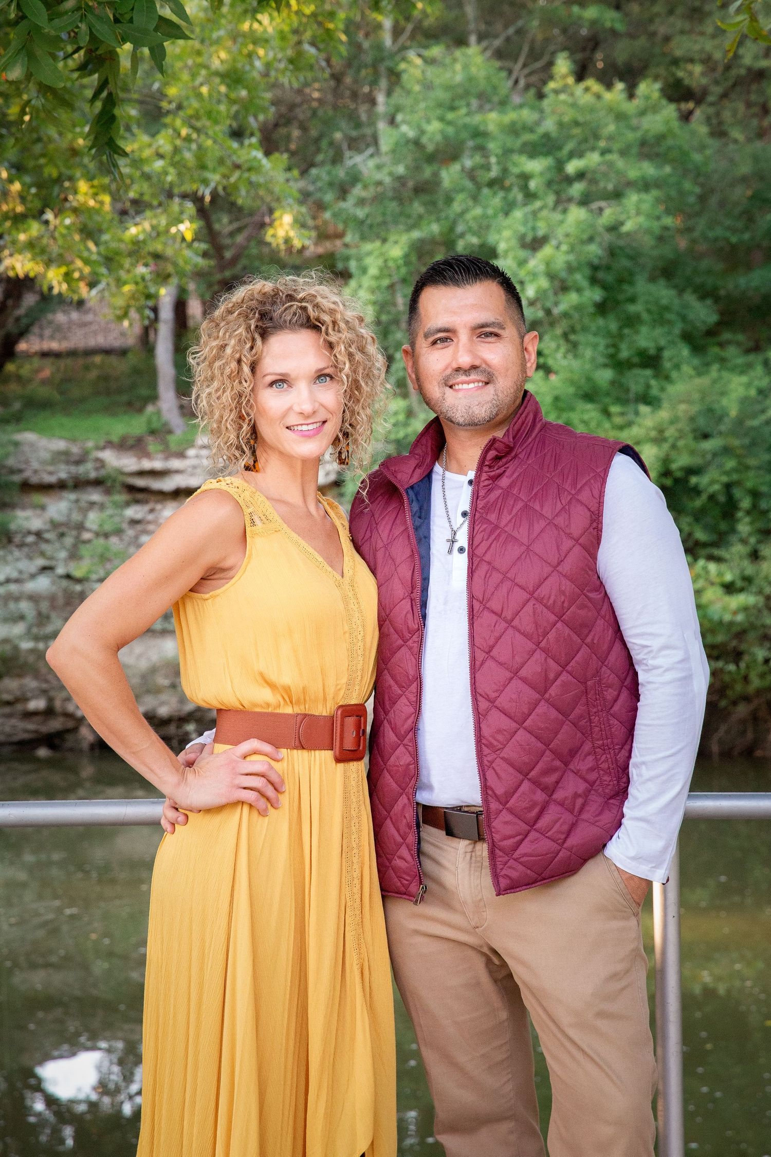 A couple poses by a pond, with her in a yellow sundress and him wearing a burgundy quilted vest.