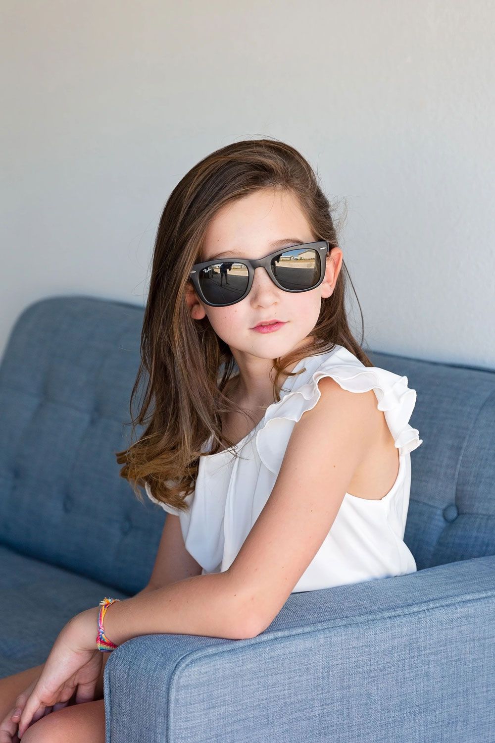 Child wearing sunglasses poses playfully on blue couch in white ruffled shirt.