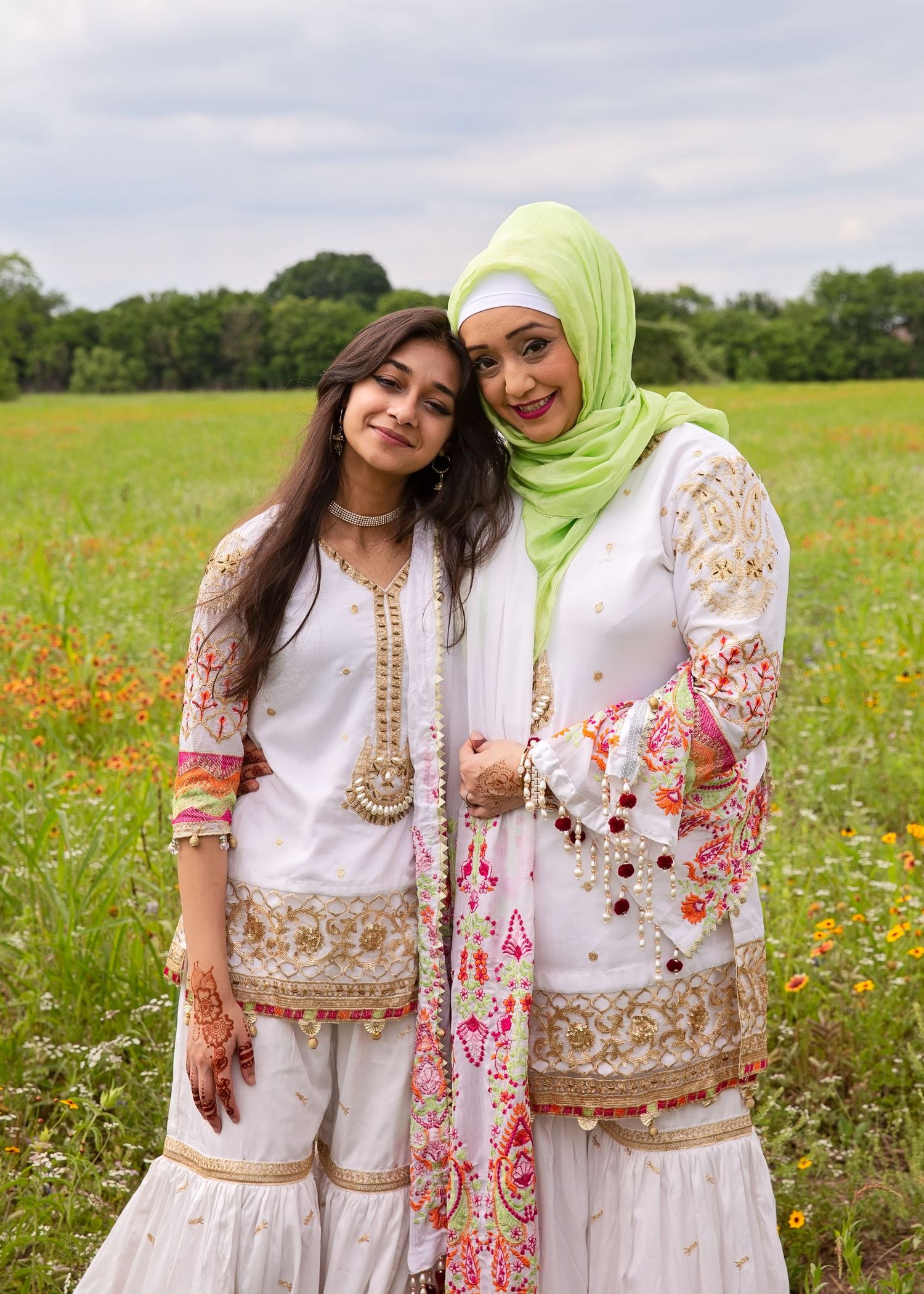 Two friends in traditional embroidered white outfits pose together in a wildflower meadow.