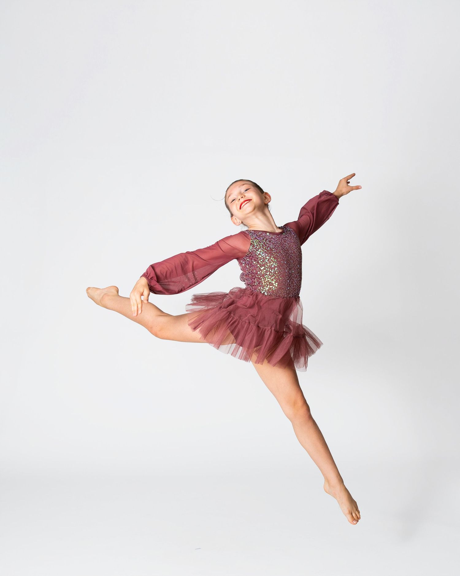 Dancer in burgundy tulle dress performing graceful leap against white backdrop.