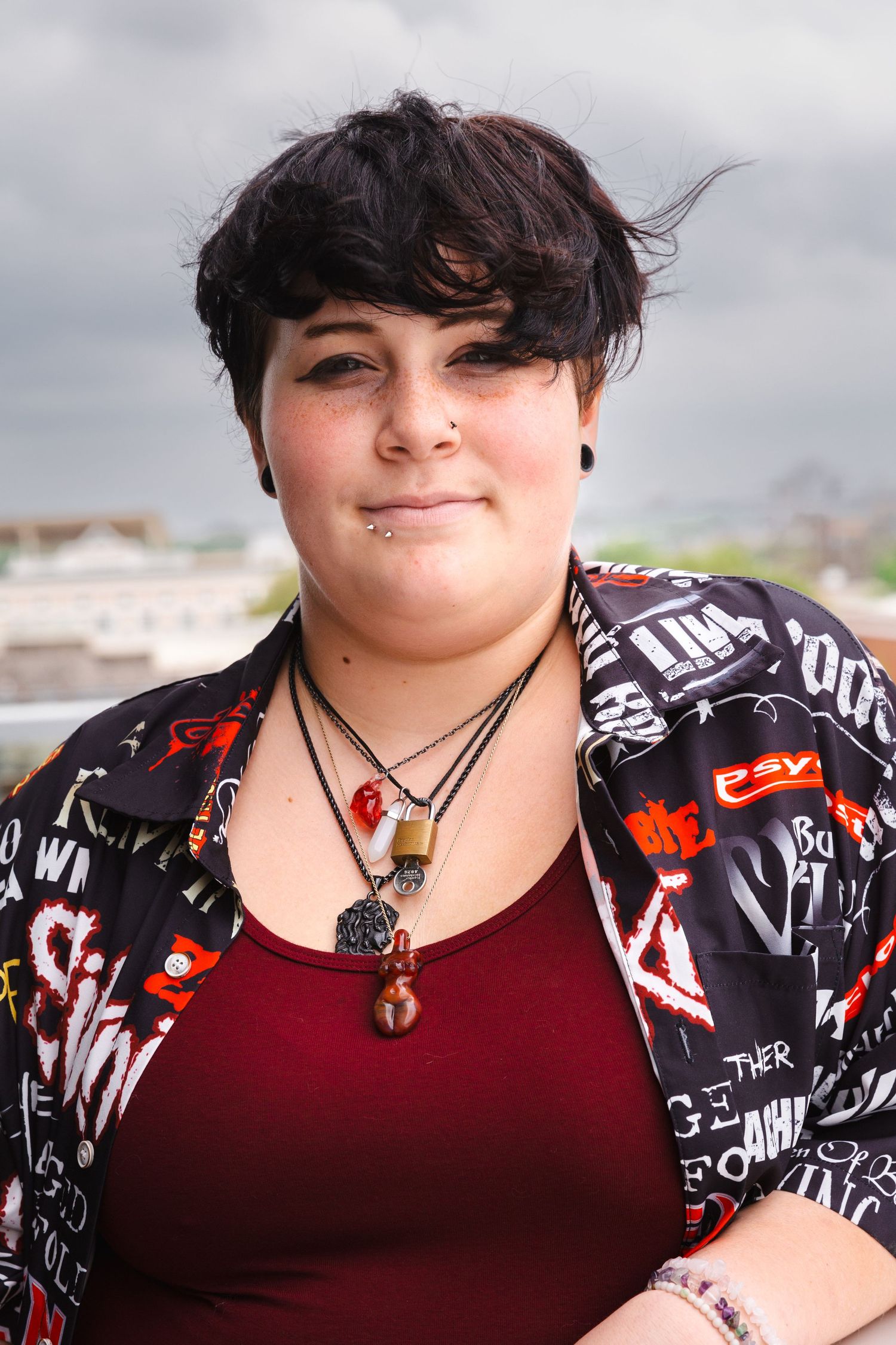 High school senior Portrait of person with short dark hair wearing graphic jacket and multiple necklaces.
