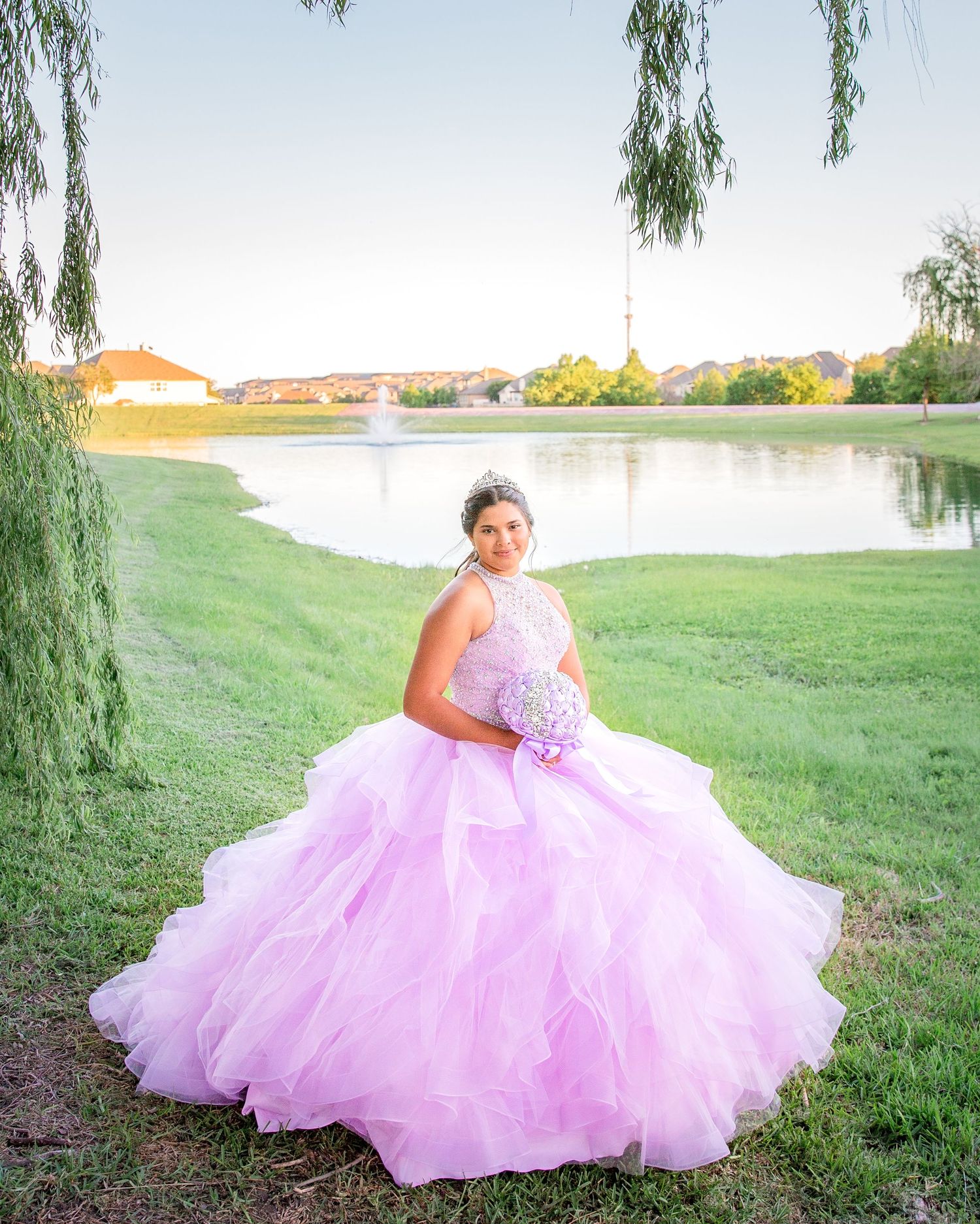 Dancer in flowing pink quinceañera gown poses by lake at sunset with willow trees.