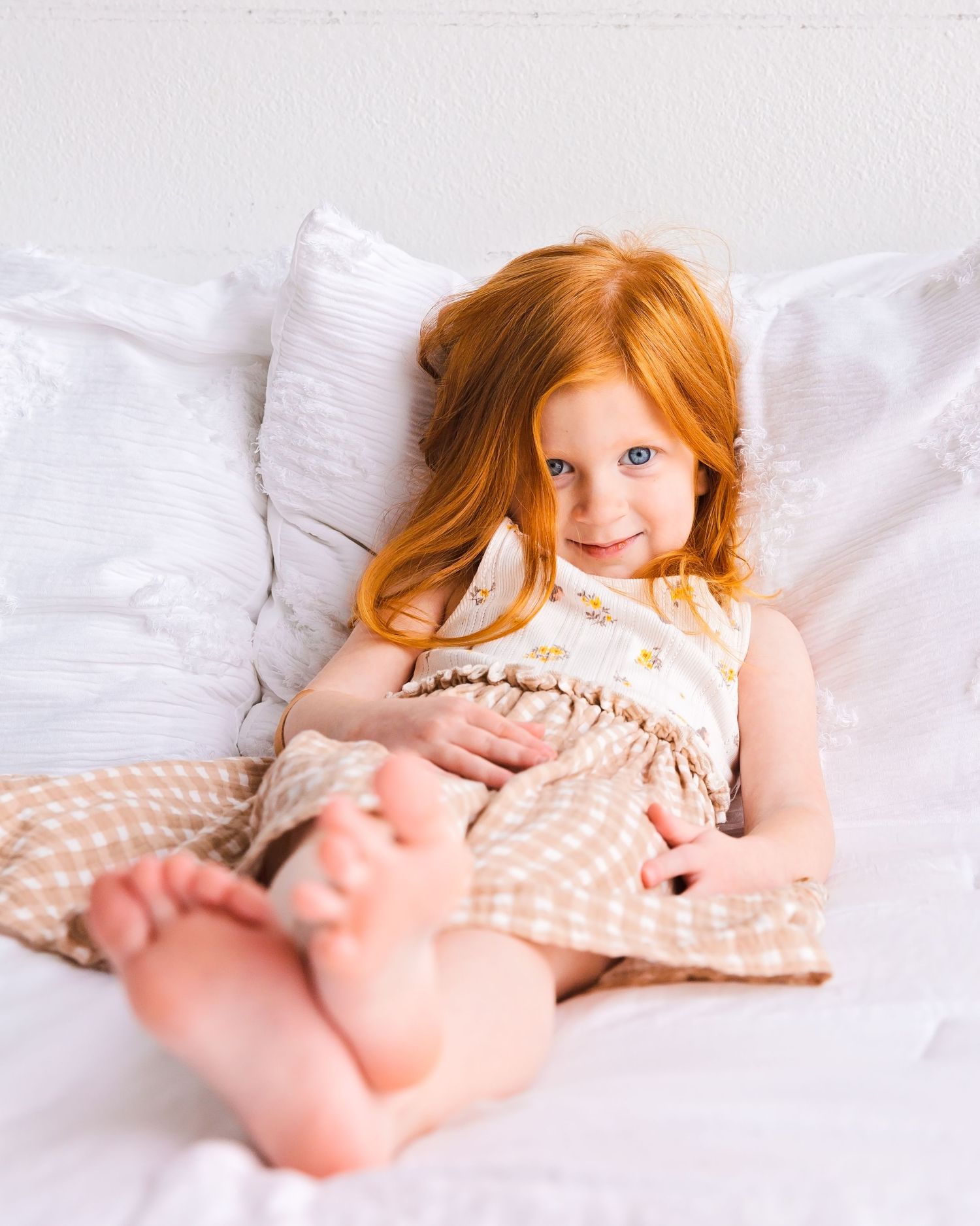 Young child with red hair lying on white bedding in a cream colored dress. Family portrait photography Round Rock, TX.