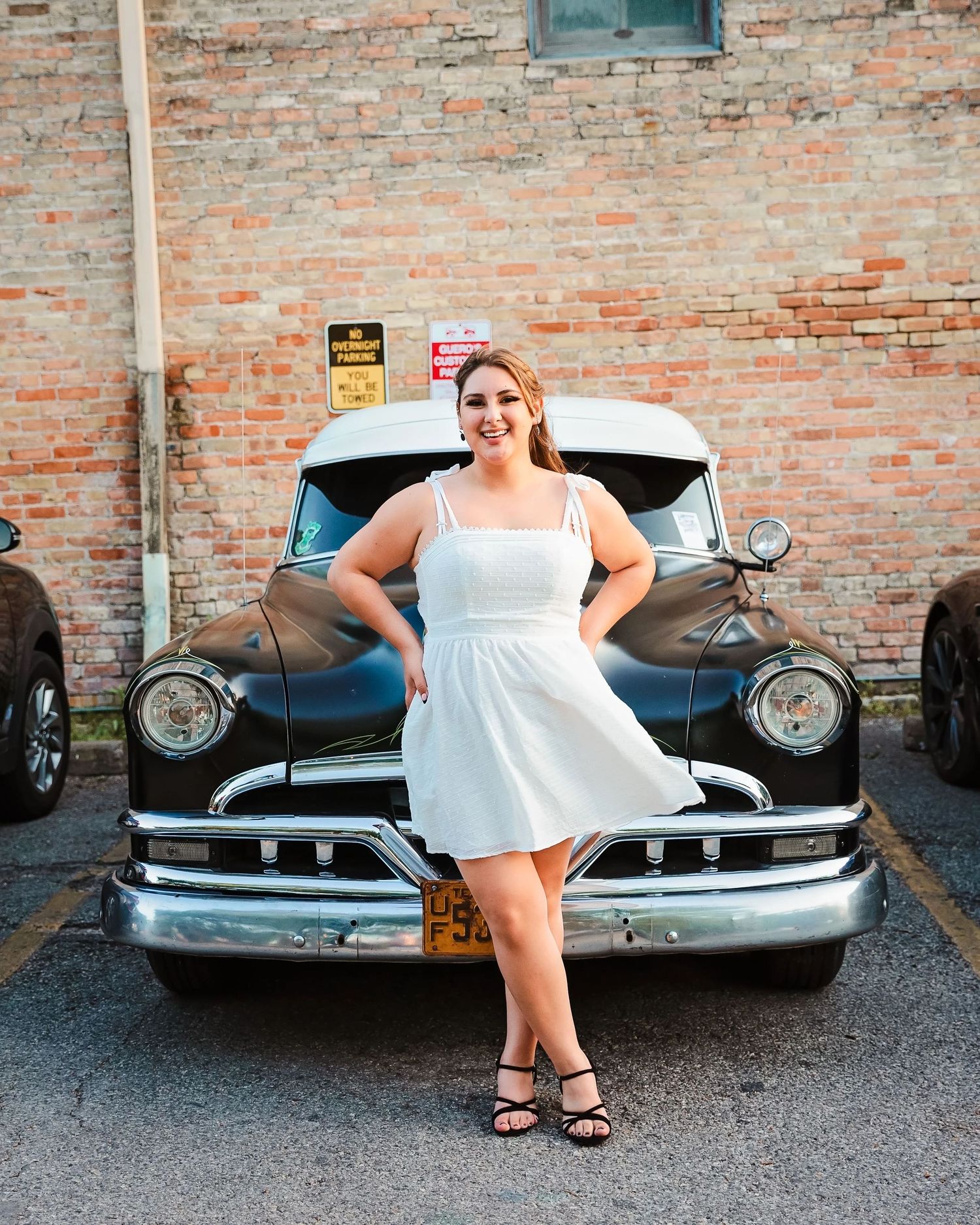 High school senior portrait. Woman in white dress poses with vintage black car against brick wall. Car show on S. Congress St in Austin, TX