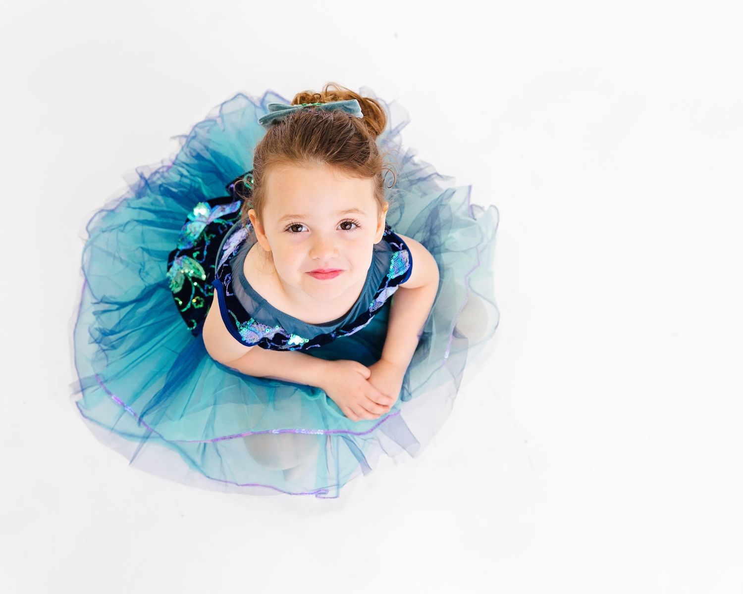 Overhead view of dancer in blue tulle dress sitting on white floor.