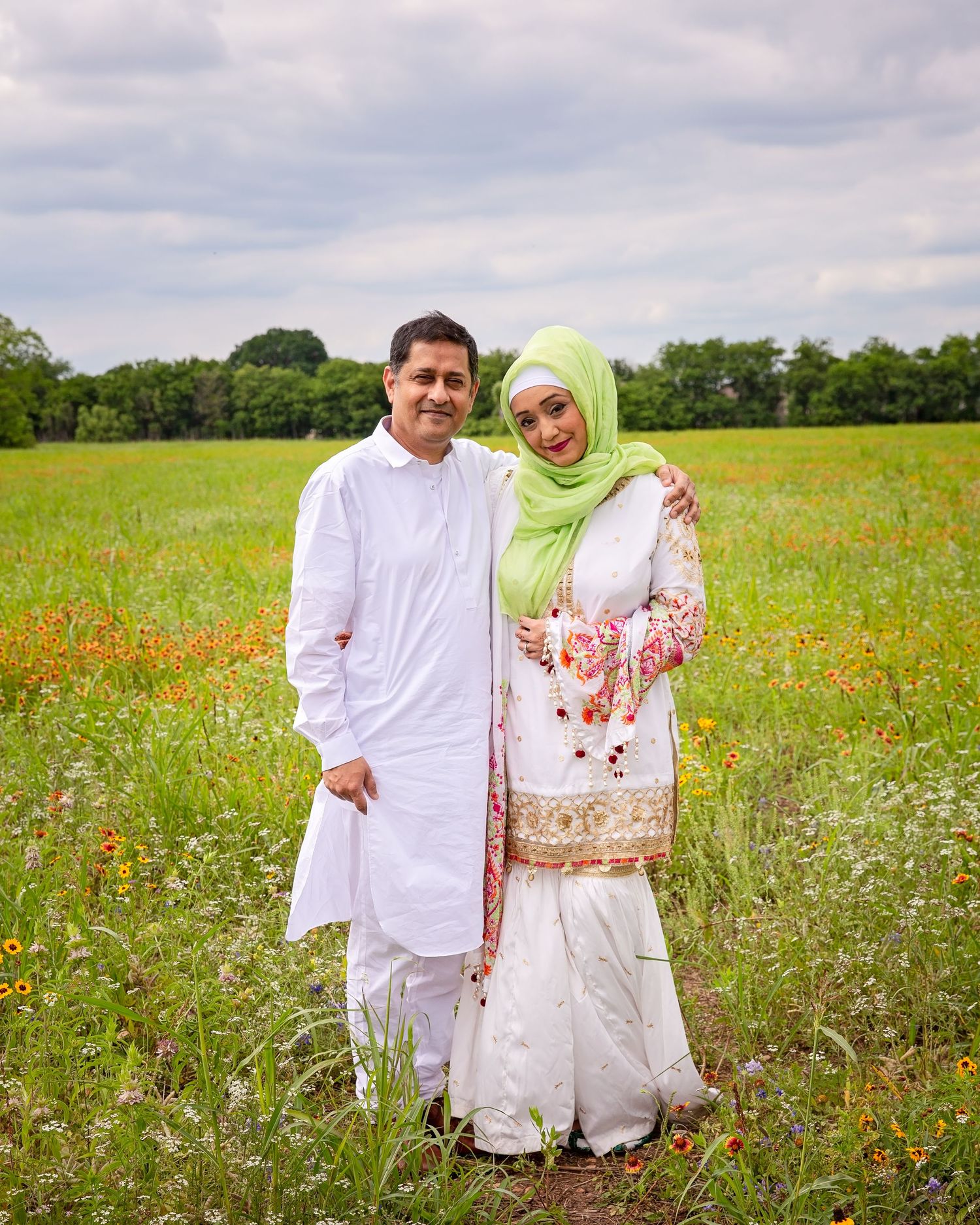 A couple in traditional white and floral attire poses together in a wildflower meadow under cloudy skies.