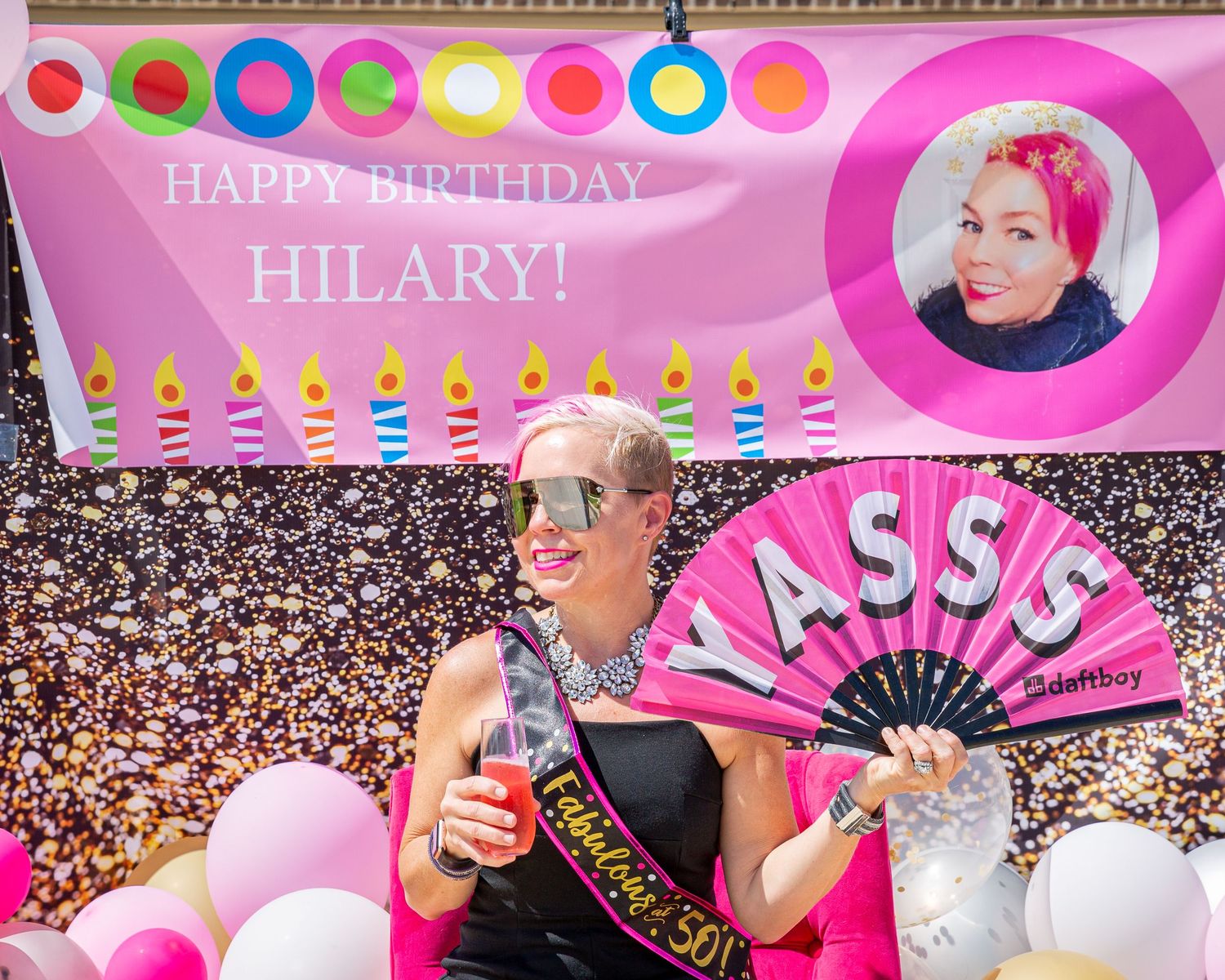 Birthday celebration with pink decorations, balloons and a festive fan against a crowded background.